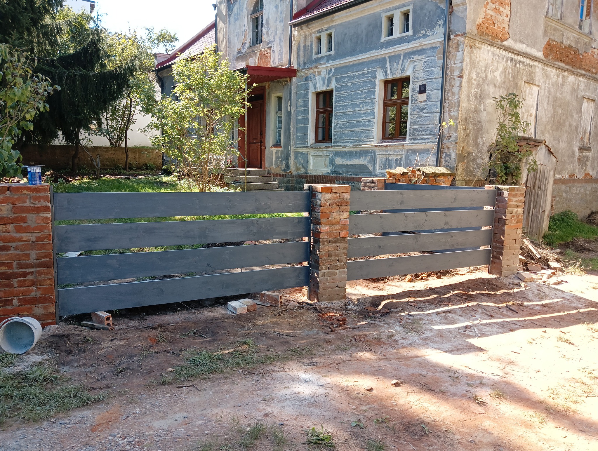 Świeżo pomalowane, poziome drewniane przęsła ogrodzenia z ceglanymi słupkami na tle odrapanego, dwupiętrowego budynku z czerwonym dachem i drewnianymi oknami.