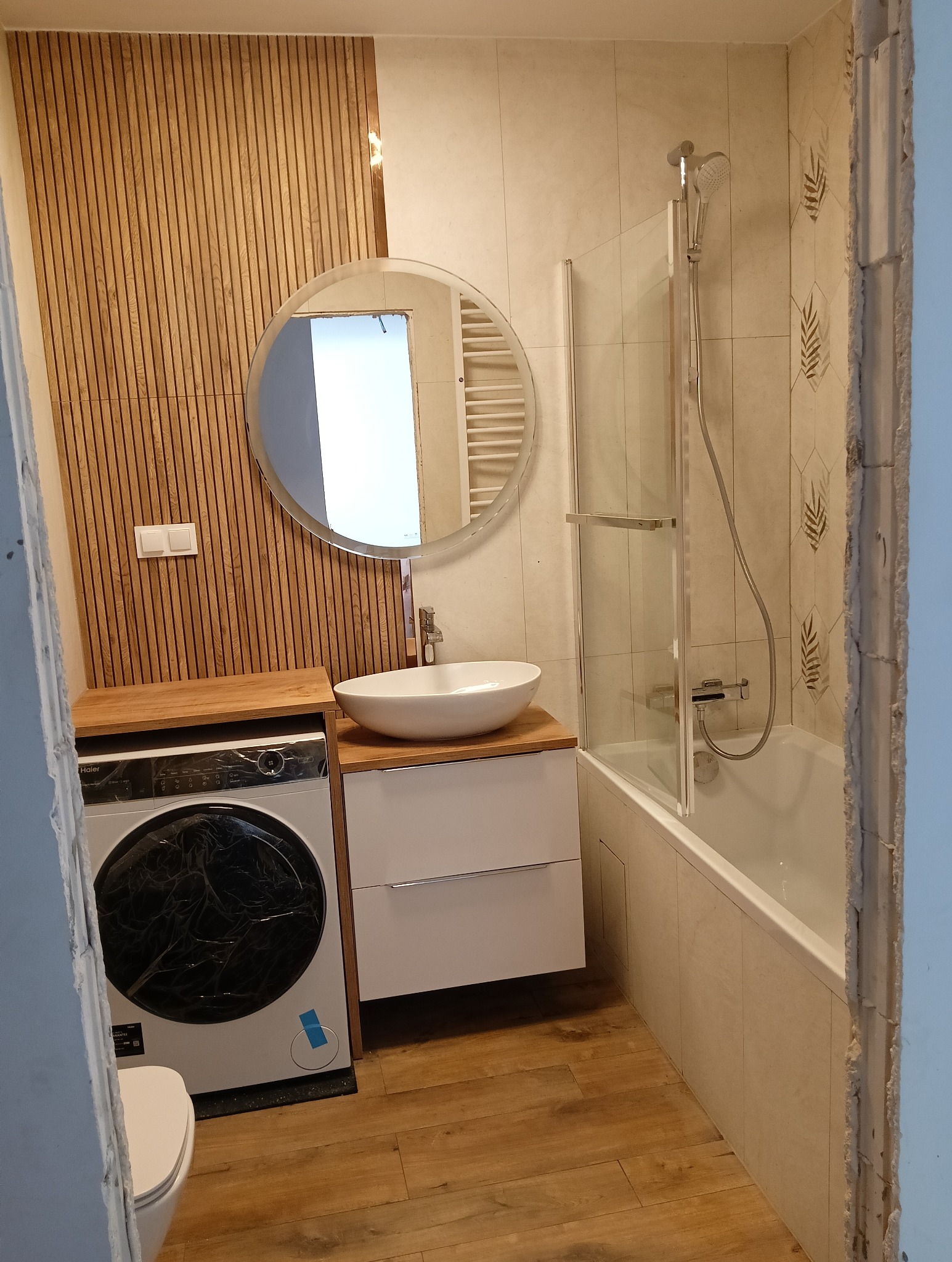 Nowoczesna łazienka w trakcie remontu: drewniana ściana z listew, okrągłe lustro nad umywalką na szafce, pralka z folią ochronną i wanna za szklanym parawanem.