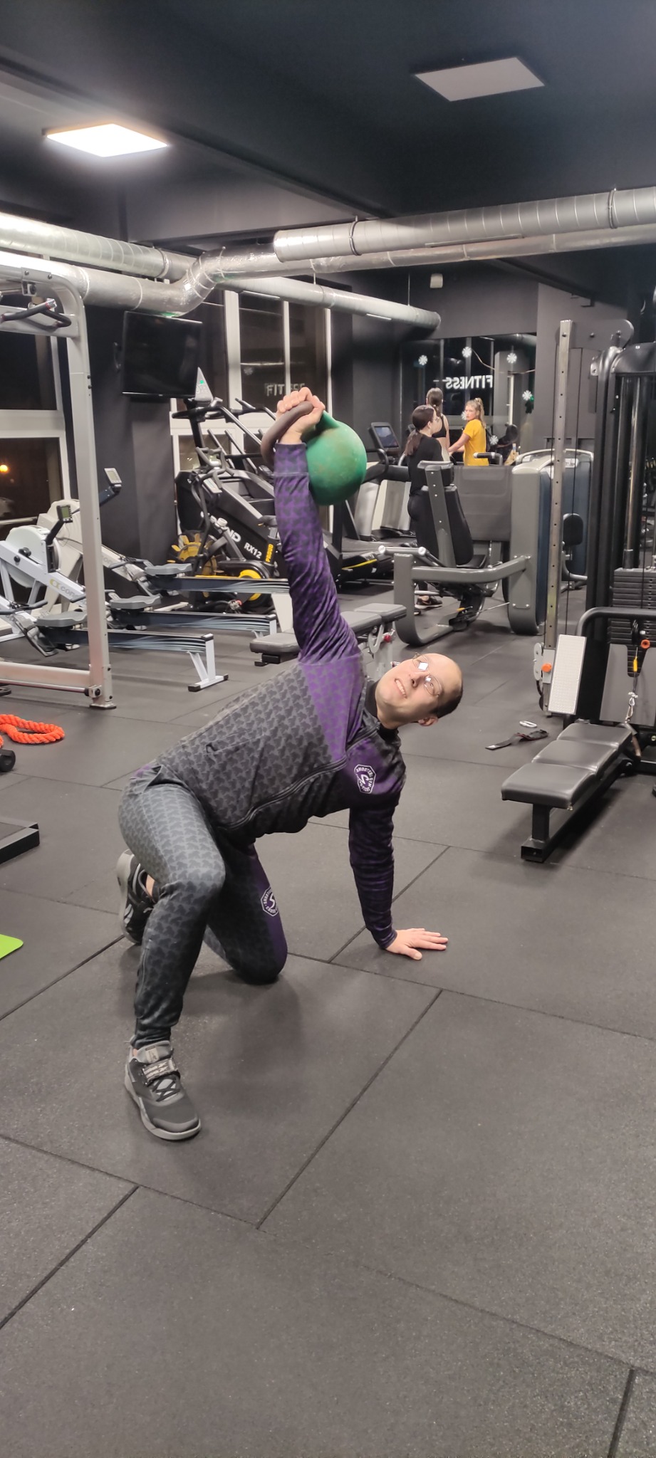 Uśmiechnięty mężczyzna w sportowym stroju, klęcząc na podłodze w siłowni, unosi zielony kettlebell nad głową, wykonując ćwiczenie w otoczeniu sprzętu do ćwiczeń.