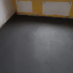 Świeżo wylana, szara posadzka jastrychowa w pomieszczeniu z żółtymi ścianami, widoczne ślady po malowaniu i gniazdka elektryczne.