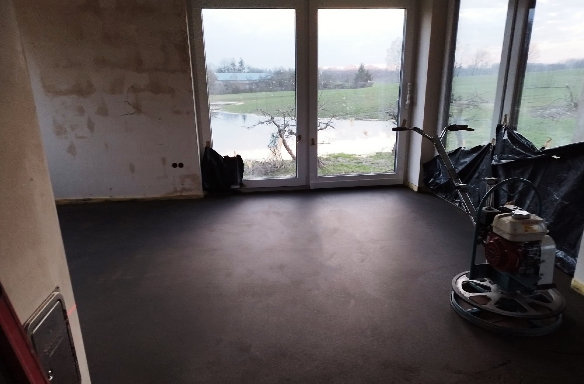 Wnętrze w trakcie wylewania posadzki jastrychowej, widoczna zacieraczka mechaniczna do betonu oraz surowe ściany i okno z widokiem na zewnątrz.