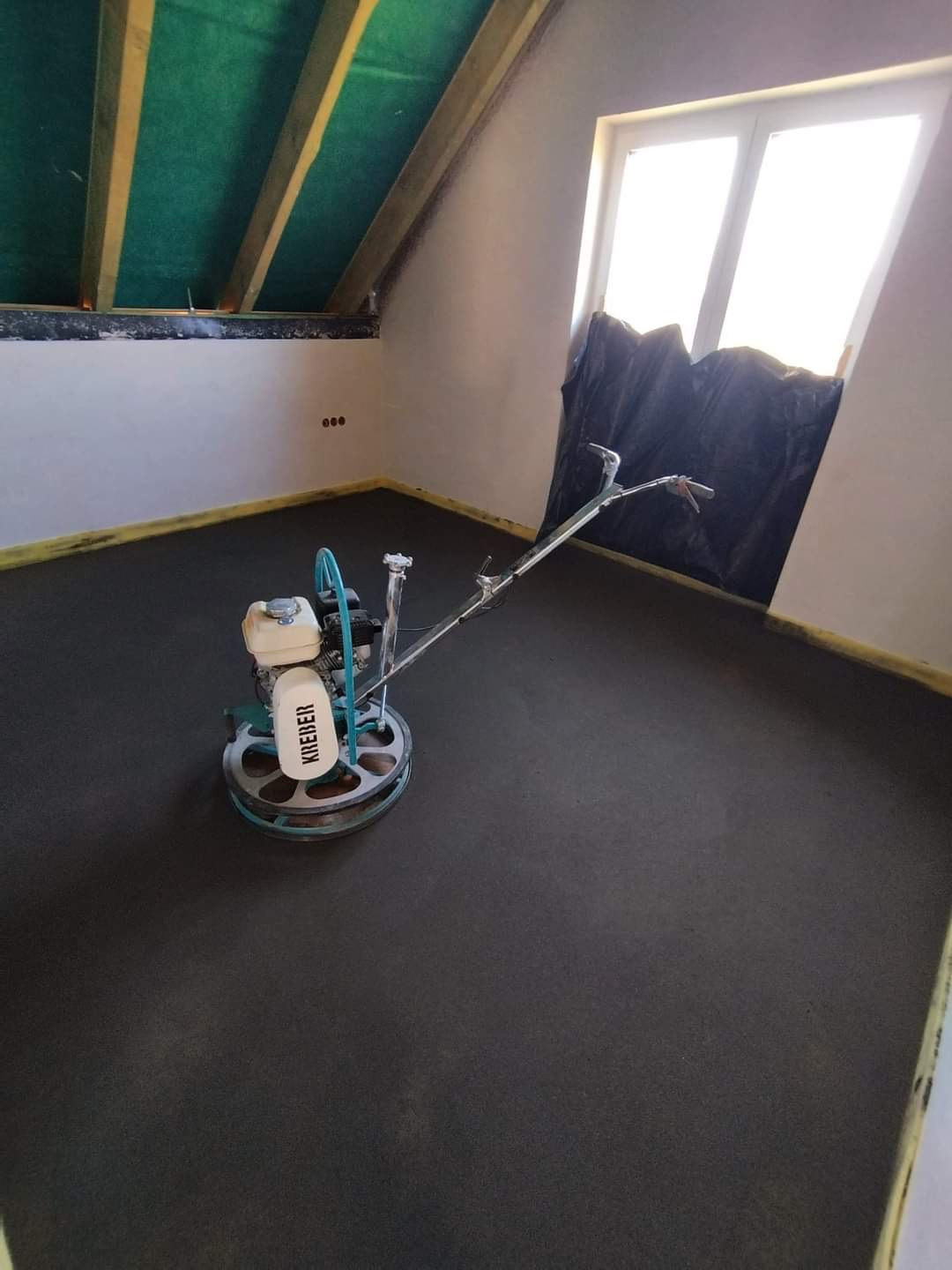 Zacieraczka mechaniczna marki Kreder na świeżo wylanym jastrychu w pomieszczeniu na poddaszu ze skosami i oknem zasłoniętym folią.