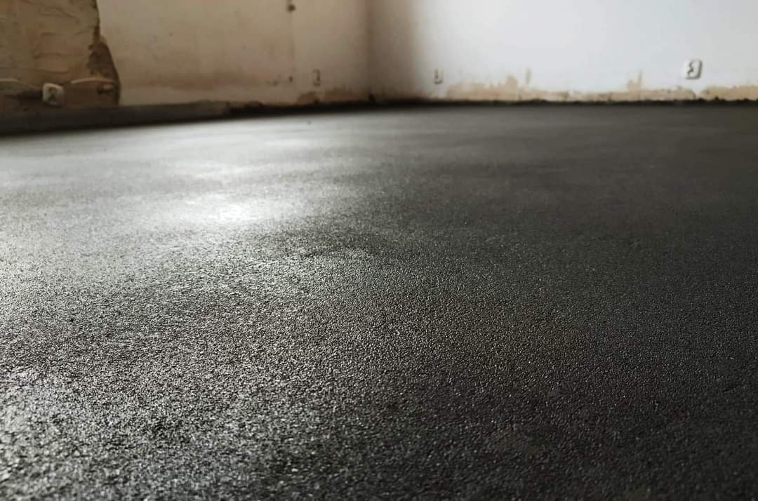 Świeżo wylana, ciemna posadzka jastrychowa z widoczną fakturą, w rogu pomieszczenia z surowymi ścianami.