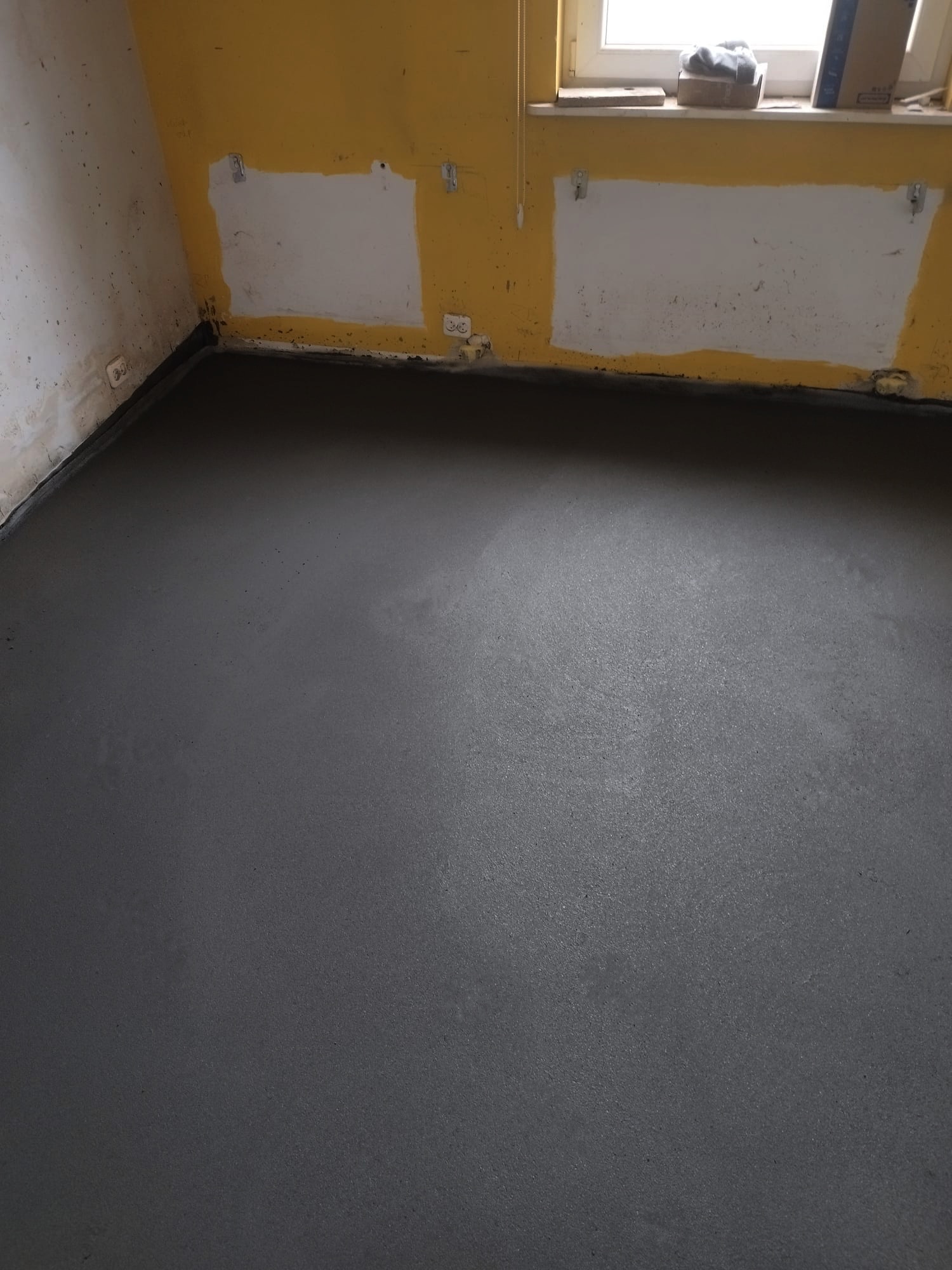 Świeżo wylana, szara posadzka jastrychowa w pomieszczeniu z żółtymi ścianami, widoczne ślady po malowaniu i gniazdka elektryczne.