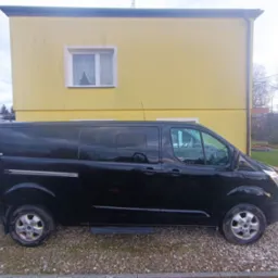 Czarny, długi bus Ford Transit na żwirowym podłożu, na tle żółtego budynku z jednym oknem i zielonego trawnika.