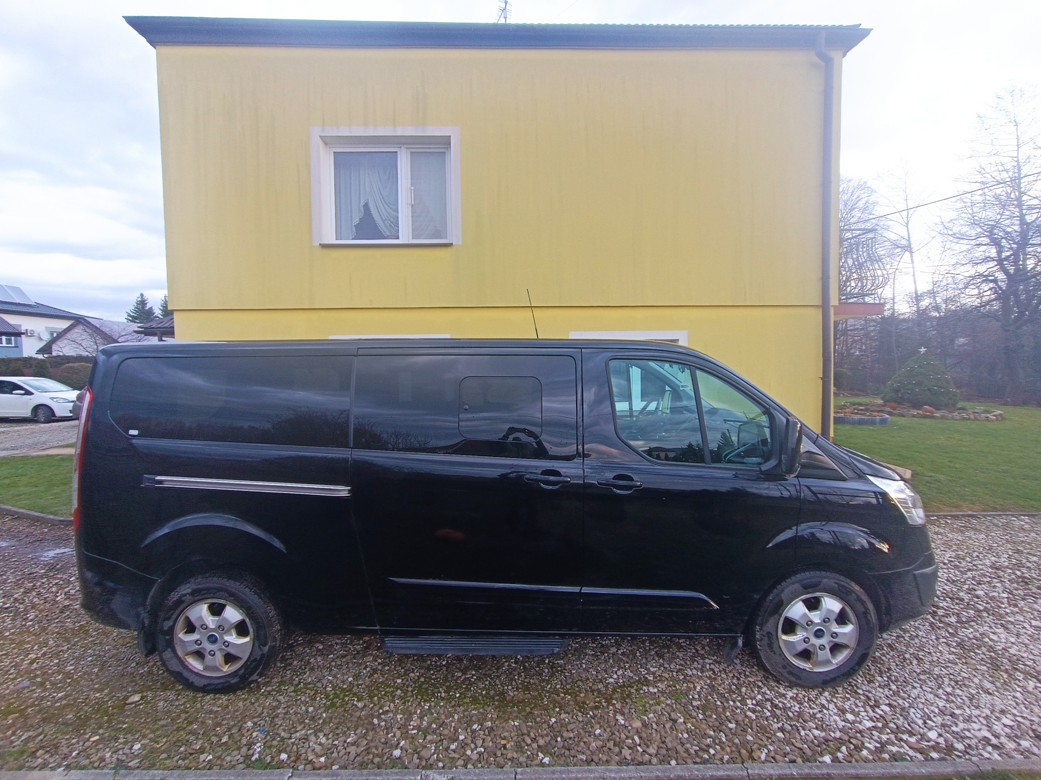 Czarny, długi bus Ford Transit na żwirowym podłożu, na tle żółtego budynku z jednym oknem i zielonego trawnika.