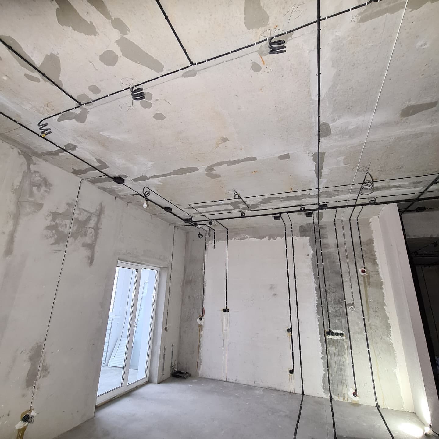 Surowy stan wykończenia wnętrza z widoczną instalacją elektryczną: czarne przewody poprowadzone po suficie i ścianach, przygotowane punkty oświetleniowe i gniazdka, ściany w stanie deweloperskim.