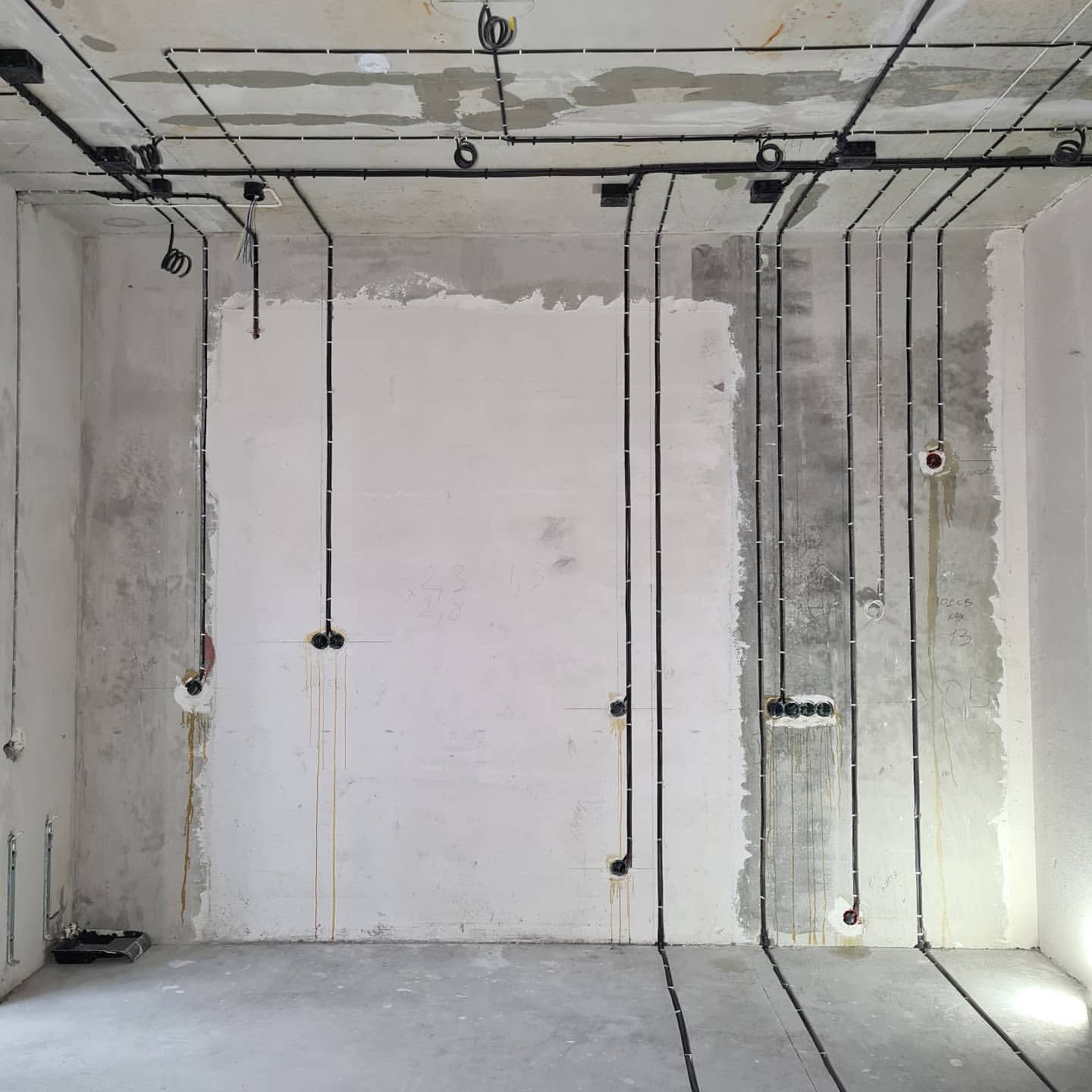 Surowy stan: czarne przewody elektryczne poprowadzone po betonowym suficie i ścianach, widoczne puszki instalacyjne, ślady po wierceniu, w tle fragment wałka malarskiego.