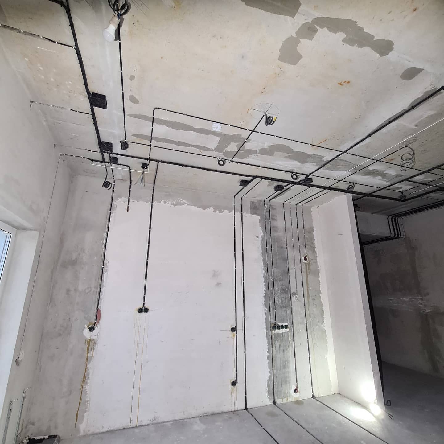 Surowy stan wnętrza z widocznymi, czarnymi przewodami elektrycznymi rozprowadzonymi po ścianach i suficie, przygotowanymi do dalszych prac wykończeniowych.