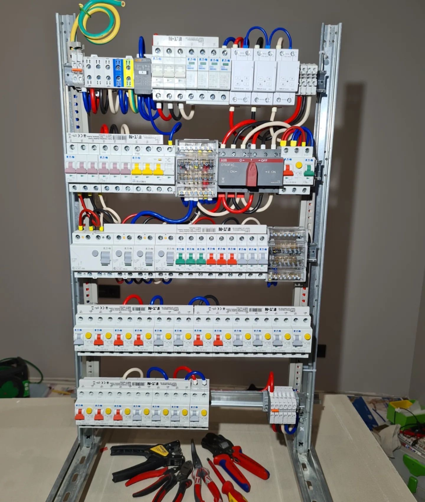 Rozdzielnica elektryczna z zainstalowanymi bezpiecznikami, wyłącznikami i przewodami w kolorach niebieskim, czerwonym, żółtym i czarnym, wraz z narzędziami elektrycznymi ułożonymi na stole poniżej.