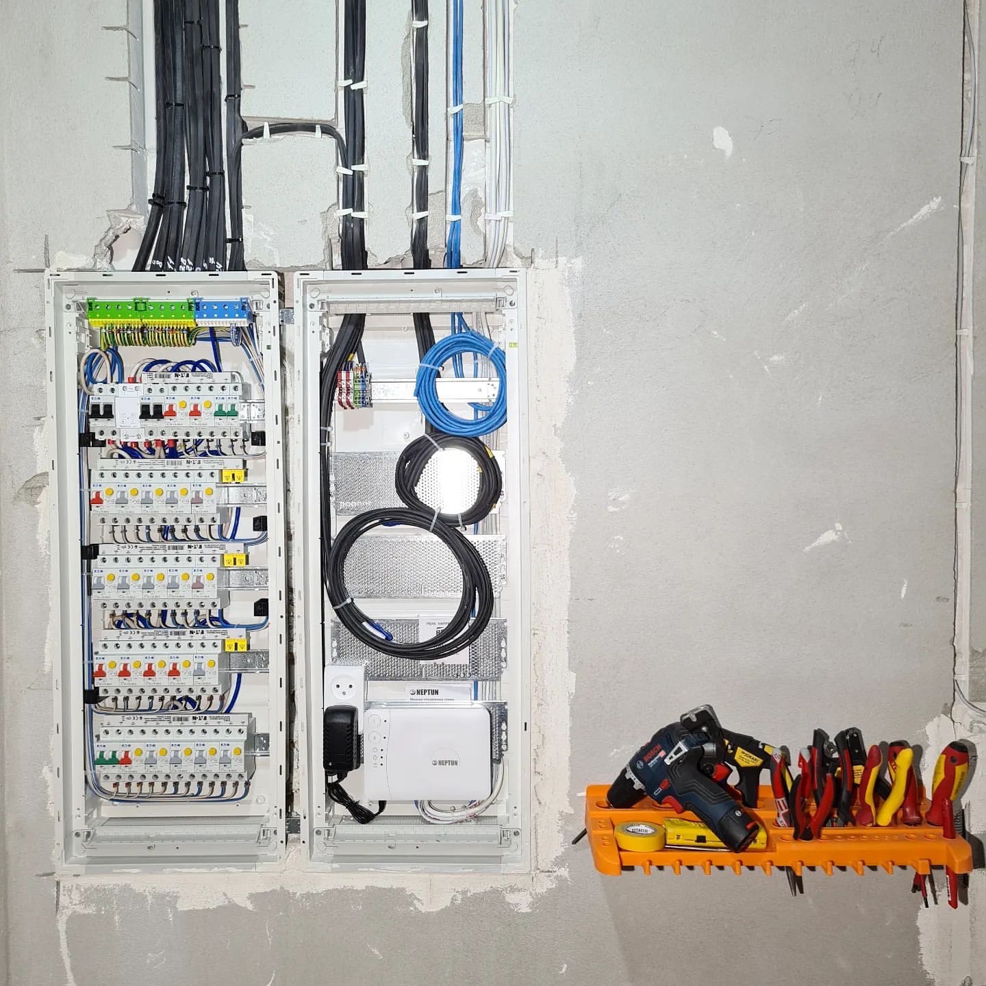 Dwa otwarte panele elektryczne z okablowaniem i bezpiecznikami, obok narzędzia na pomarańczowym wieszaku na surowej ścianie.