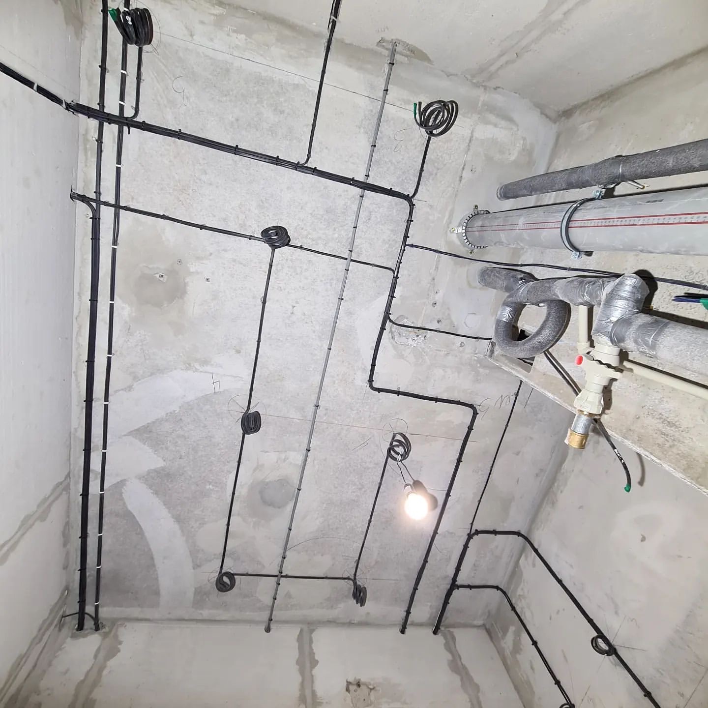 Sufit w surowym stanie z rozprowadzonymi czarnymi kablami elektrycznymi, widoczne puszki instalacyjne i tymczasowa żarówka, obok rury wentylacyjne.