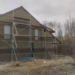 Dom jednorodzinny w trakcie prac wykończeniowych elewacji, otoczony rusztowaniem, z widocznymi niebieskimi rurami i folią ochronną na oknach, na tle pochmurnego nieba i drzew bez liści.