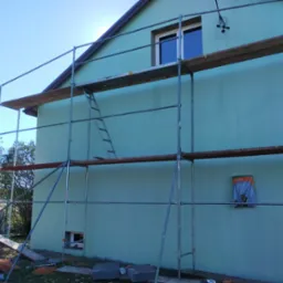 Ściana budynku w kolorze turkusowym, częściowo zasłonięta rusztowaniem. Widoczny fragment okna i wystający kabel. Na ziemi leżą materiały budowlane.