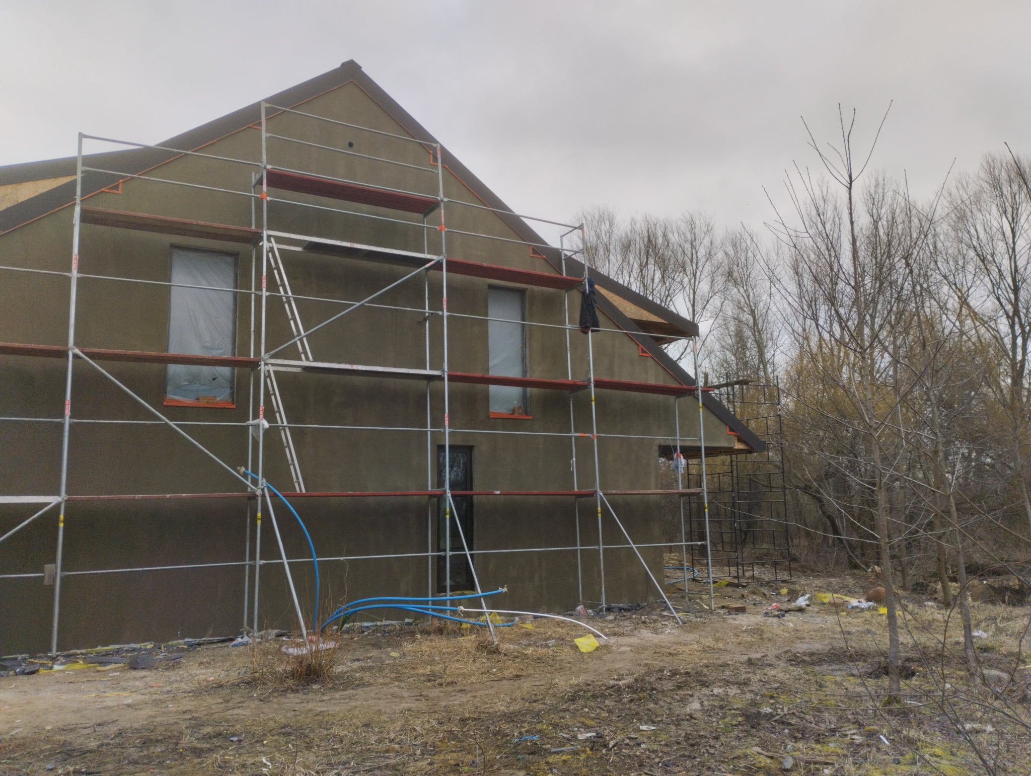 Dom jednorodzinny w trakcie prac wykończeniowych elewacji, otoczony rusztowaniem, z widocznymi niebieskimi rurami i folią ochronną na oknach, na tle pochmurnego nieba i drzew bez liści.