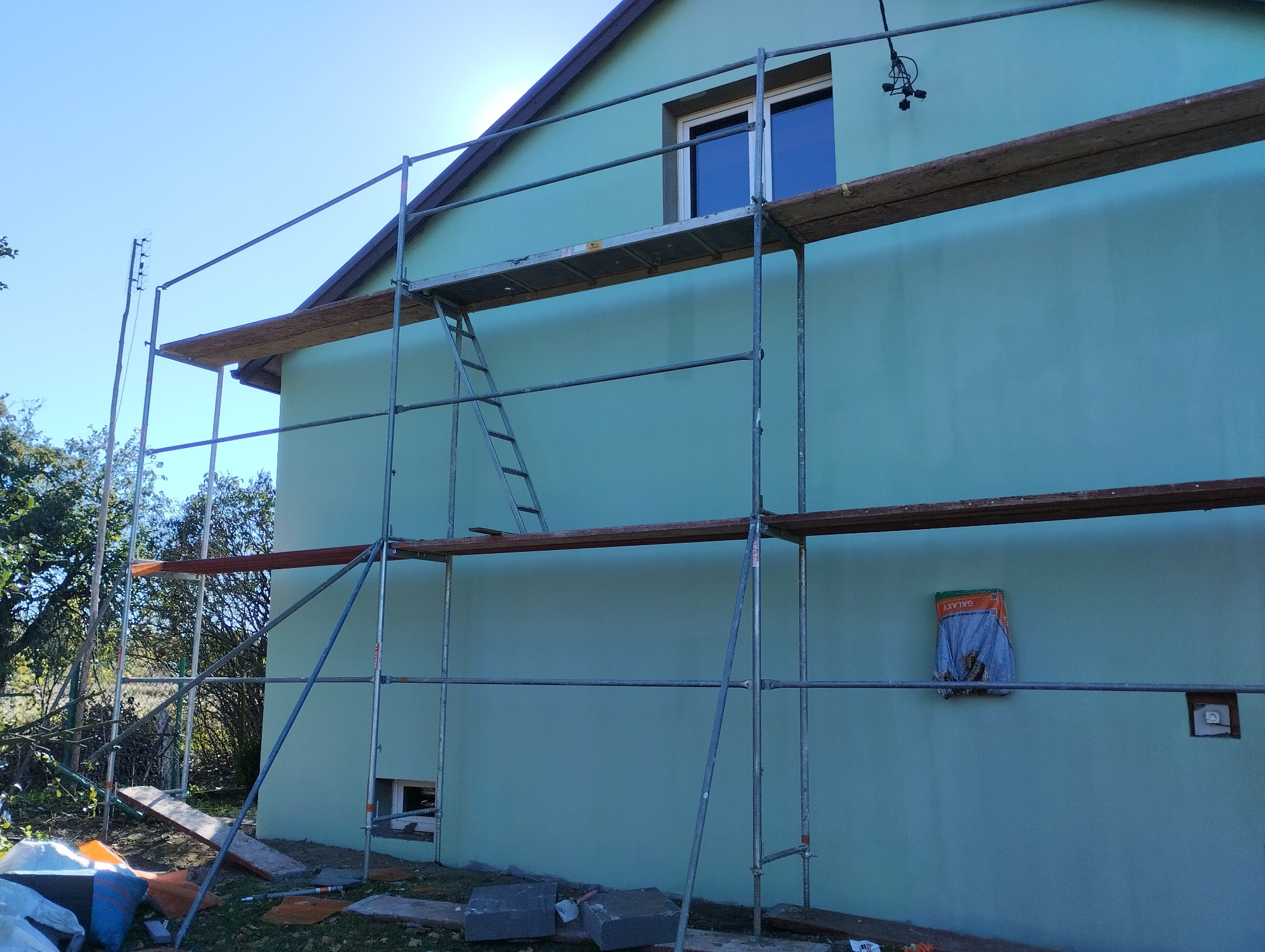 Ściana budynku w kolorze turkusowym, częściowo zasłonięta rusztowaniem. Widoczny fragment okna i wystający kabel. Na ziemi leżą materiały budowlane.