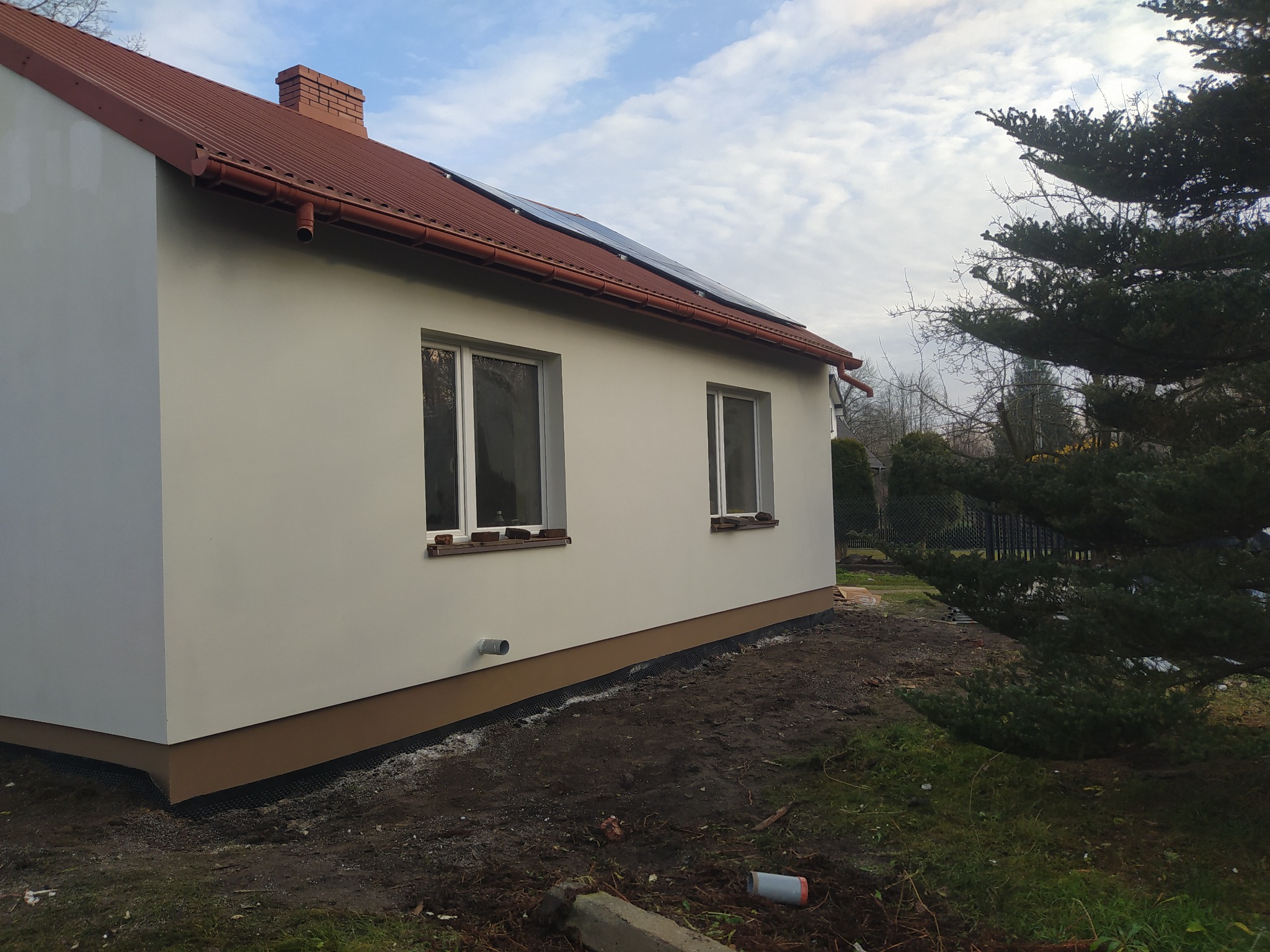 Jasny, otynkowany dom jednorodzinny z brązowym dachem i panelami słonecznymi, widziany z boku, z fragmentem ogrodu i drzew iglastych w tle.