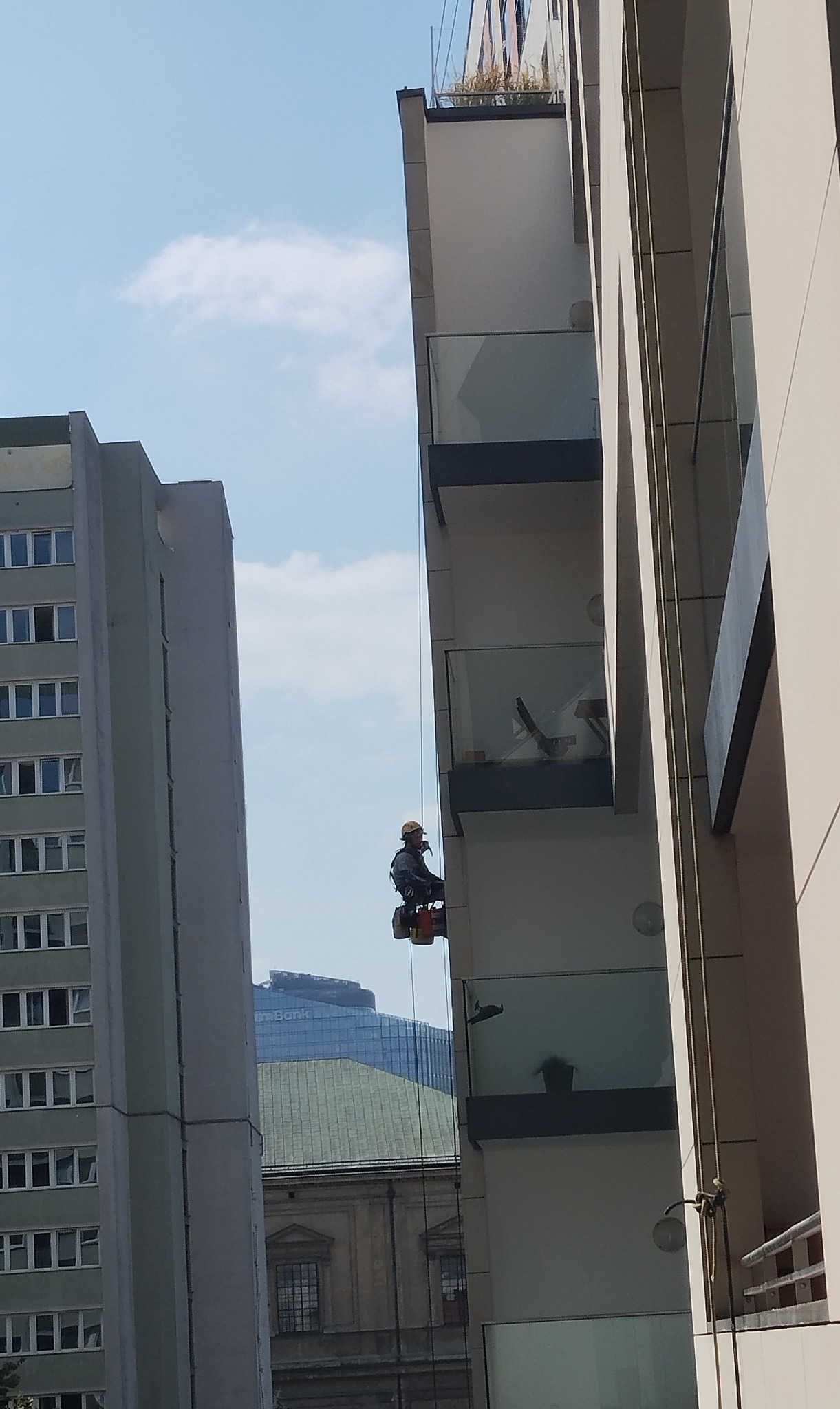 Pracownik w kasku, na linach, podczas prac na wysokości na elewacji nowoczesnego budynku z balkonami, w tle inne budynki i błękitne niebo.