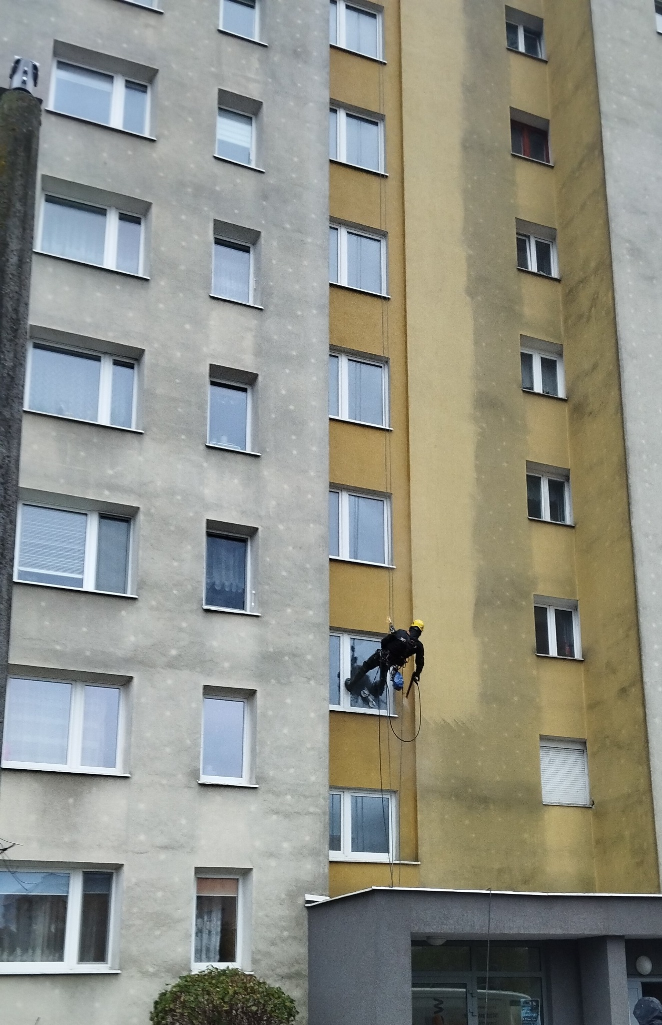Pracownik w uprzęży, w kasku, zawieszony na linach, wykonuje prace na wysokości na elewacji bloku mieszkalnego. Widoczne okna i fragmenty elewacji w kolorach szarym i żółtym.