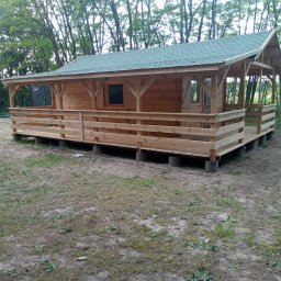 Usługi Stolarskie - Niewielki drewniany domek z zadaszonym tarasem, posadowiony na betonowych bloczkach, z zielonym dachem, otoczony drzewami i trawiastym terenem.