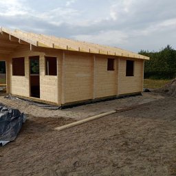 Usługi Stolarskie - Wznoszona konstrukcja domu z drewna, widoczne ściany z bali i elementy więźby dachowej, teren budowy z piaskiem i folią ochronną.