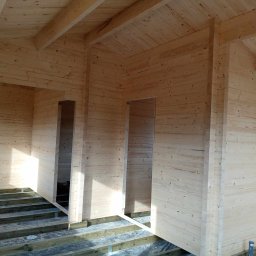 Usługi Stolarskie - Wnętrze surowego drewnianego domu w trakcie budowy, widoczne ściany z bali, drewniany strop i przygotowane podłoże pod podłogę.