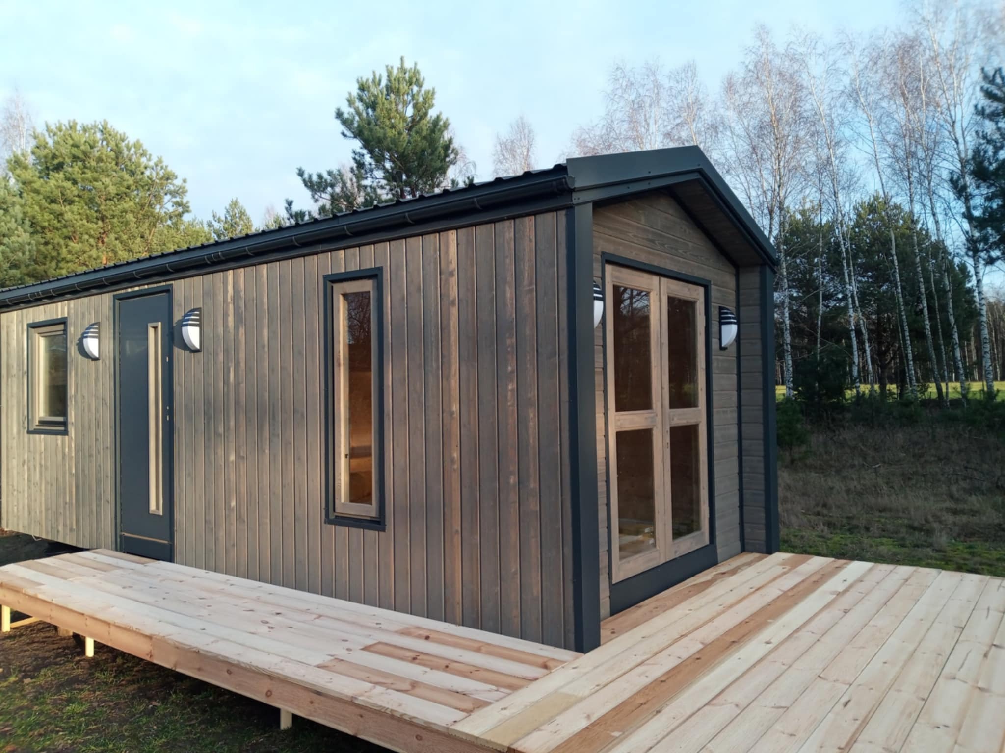 Gotowy, parterowy budynek z drewnianą elewacją i dobudowanym tarasem, otoczony zielenią i drzewami w tle.