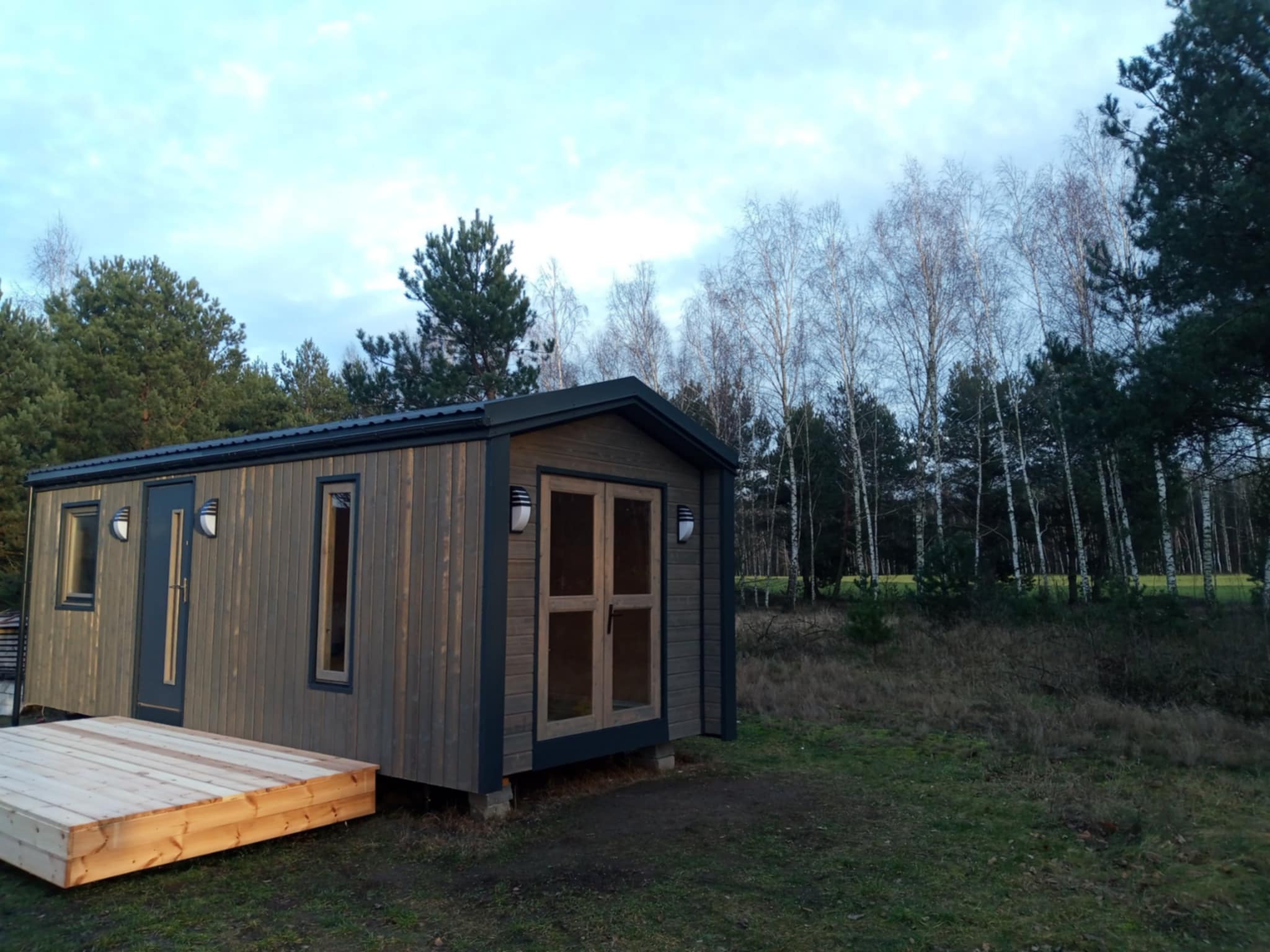 Gotowy, ciemnoszary budynek z drewna z drewnianym tarasem na trawiastym terenie, otoczony drzewami z bladym, zachmurzonym niebem w tle.