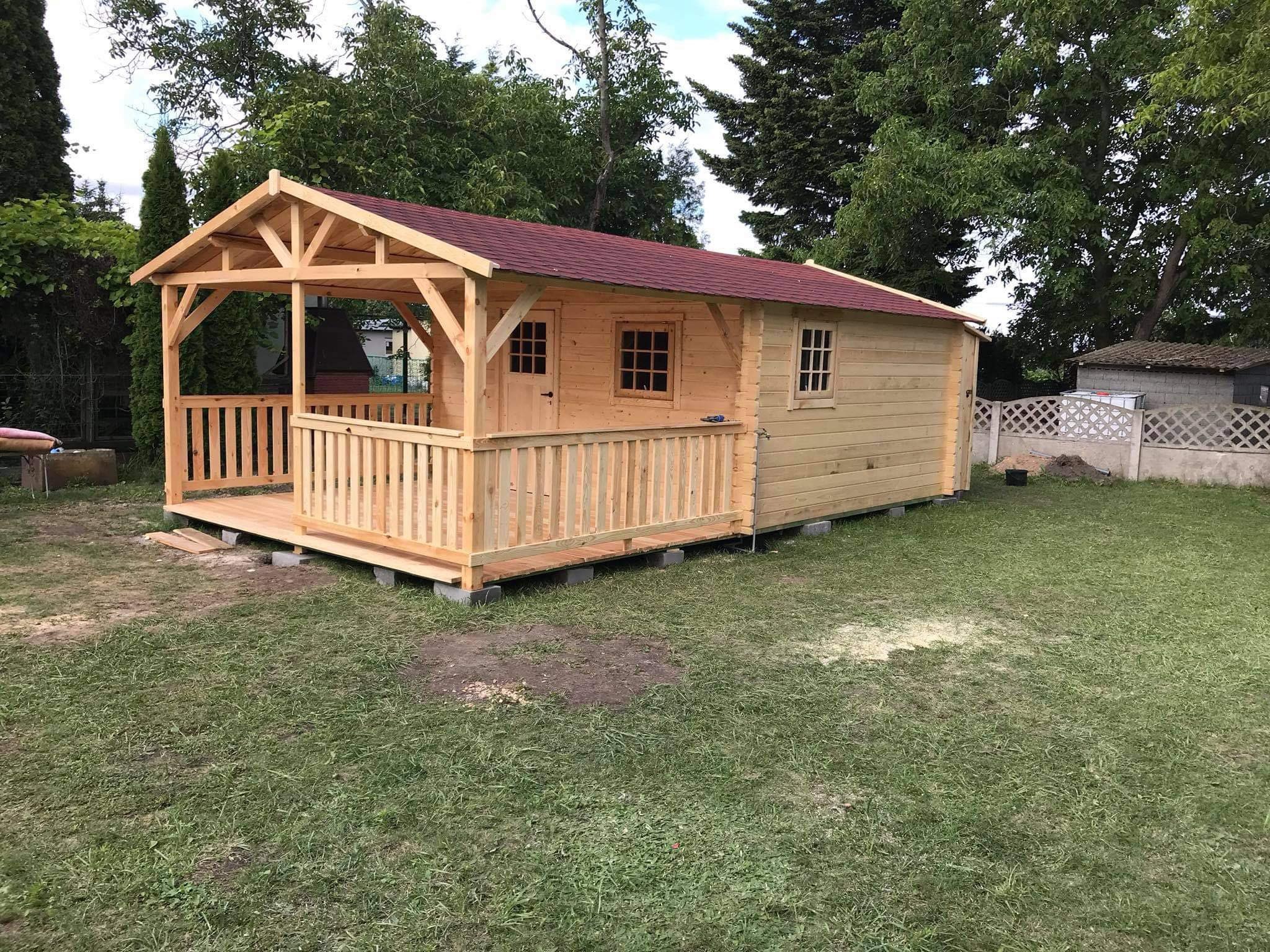 Niewielki, jasny drewniany domek z czerwonym dachem i zadaszonym tarasem, posadowiony na betonowych bloczkach na trawniku.