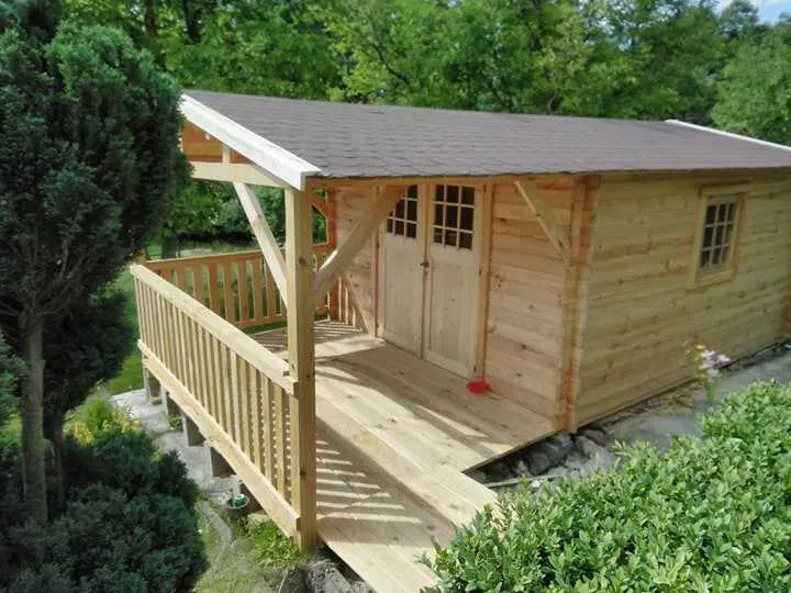 Niewielki, drewniany budynek z zadaszonym tarasem, brązowym dachem i jasnymi drzwiami z okienkami, otoczony zielenią.