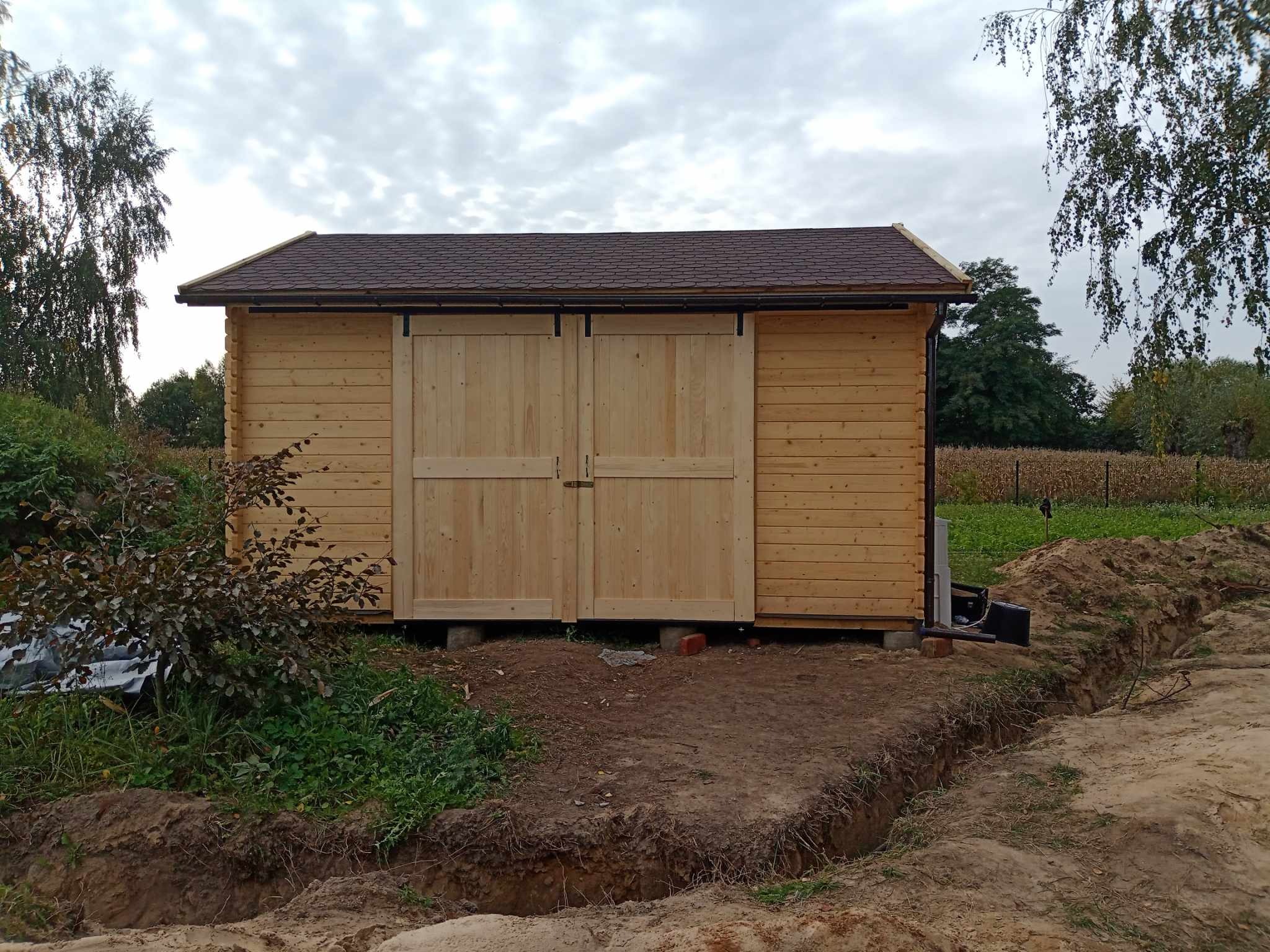 Drewniany domek z bali z przesuwnymi drzwiami, brązowym dachem z gontu bitumicznego, posadowiony na bloczkach, widoczny wykop w ziemi.