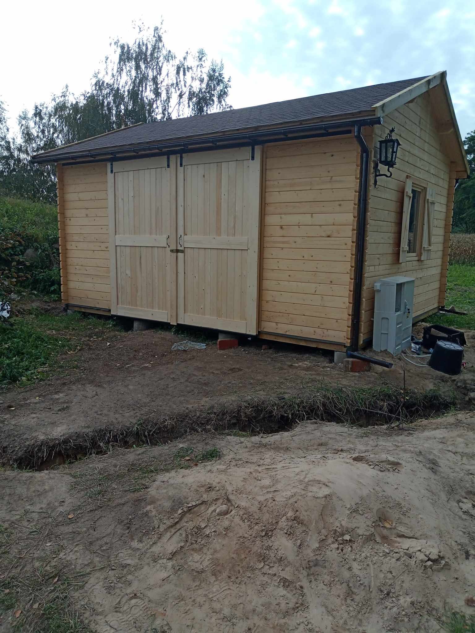 Mały, drewniany budynek z przesuwnymi drzwiami i oświetleniem zewnętrznym, posadowiony na bloczkach, z wykopem w ziemi w tle.