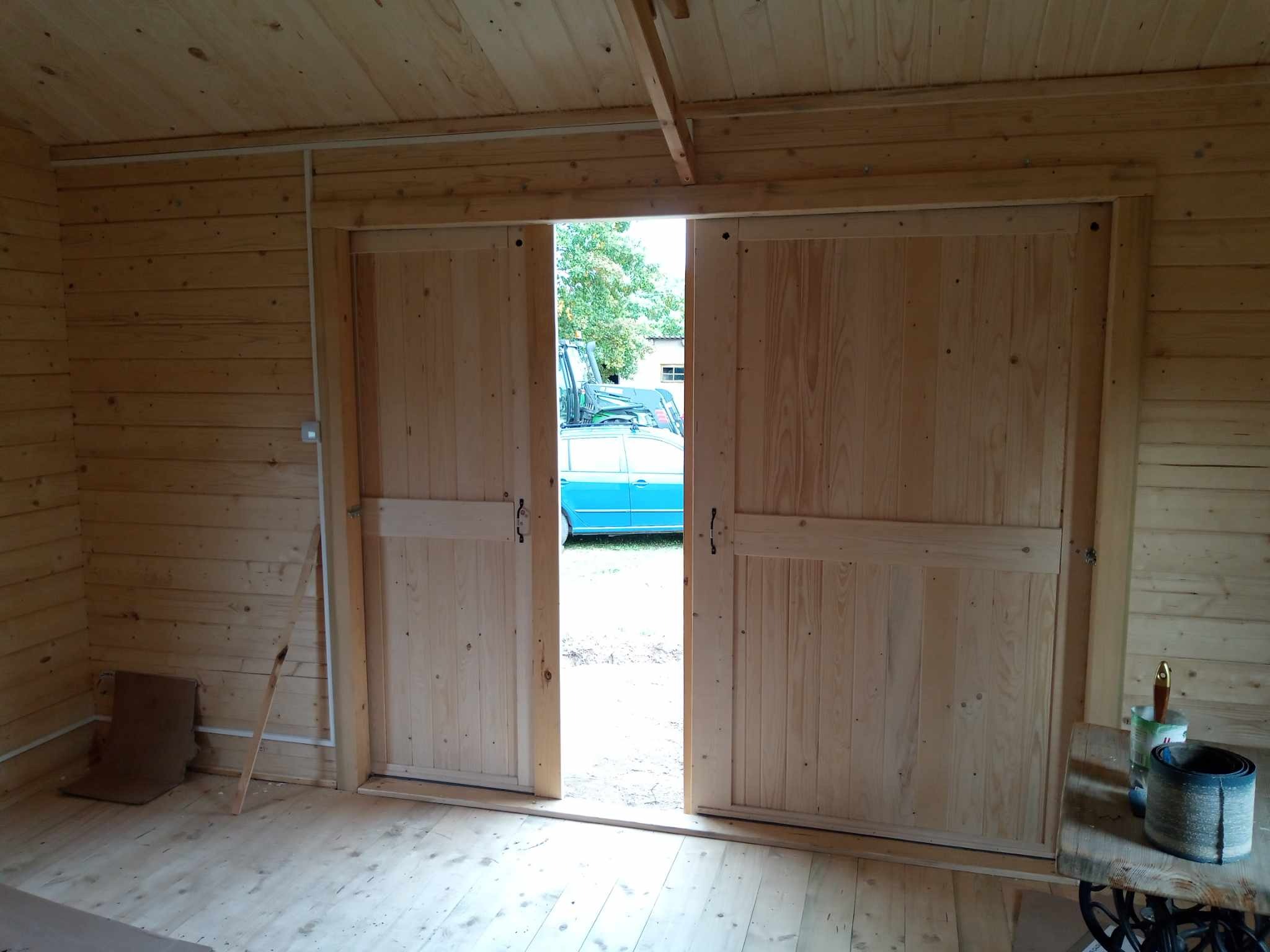 Wnętrze drewnianej konstrukcji z przesuwanymi drzwiami stodołowymi, widoczny fragment niebieskiego pojazdu na zewnątrz.