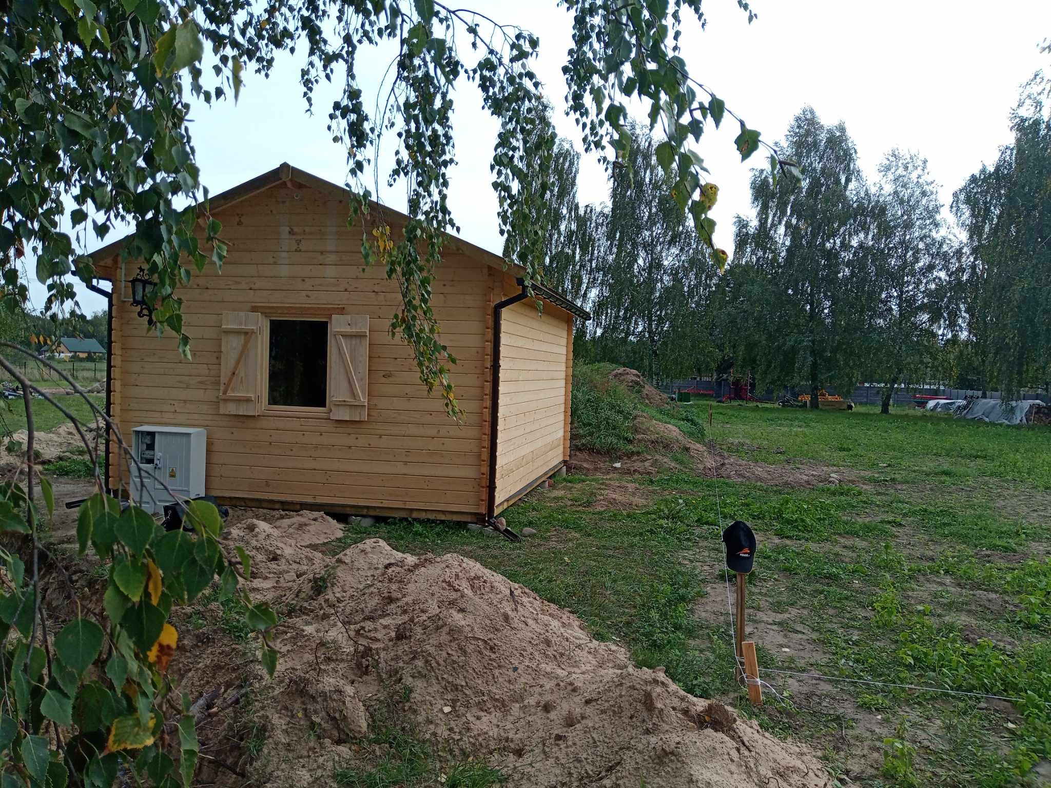 Surowy, drewniany domek z okiennicami w trakcie budowy, widoczne kopce ziemi i skrzynka elektryczna na zewnątrz, zieleń drzew w tle.
