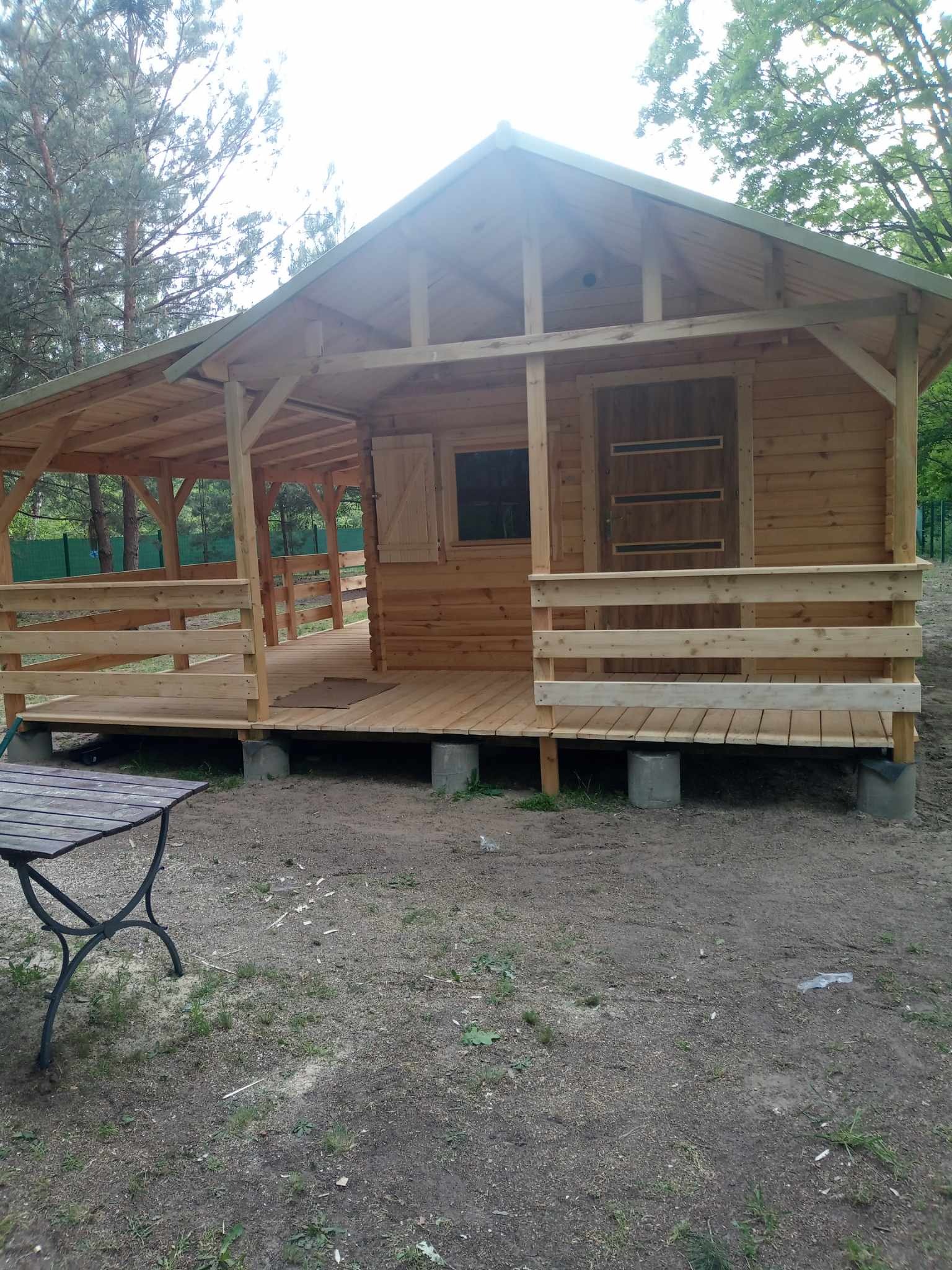 Surowy drewniany domek z zadaszonym tarasem, posadowiony na betonowych bloczkach, z widocznym stołem ogrodowym w tle.