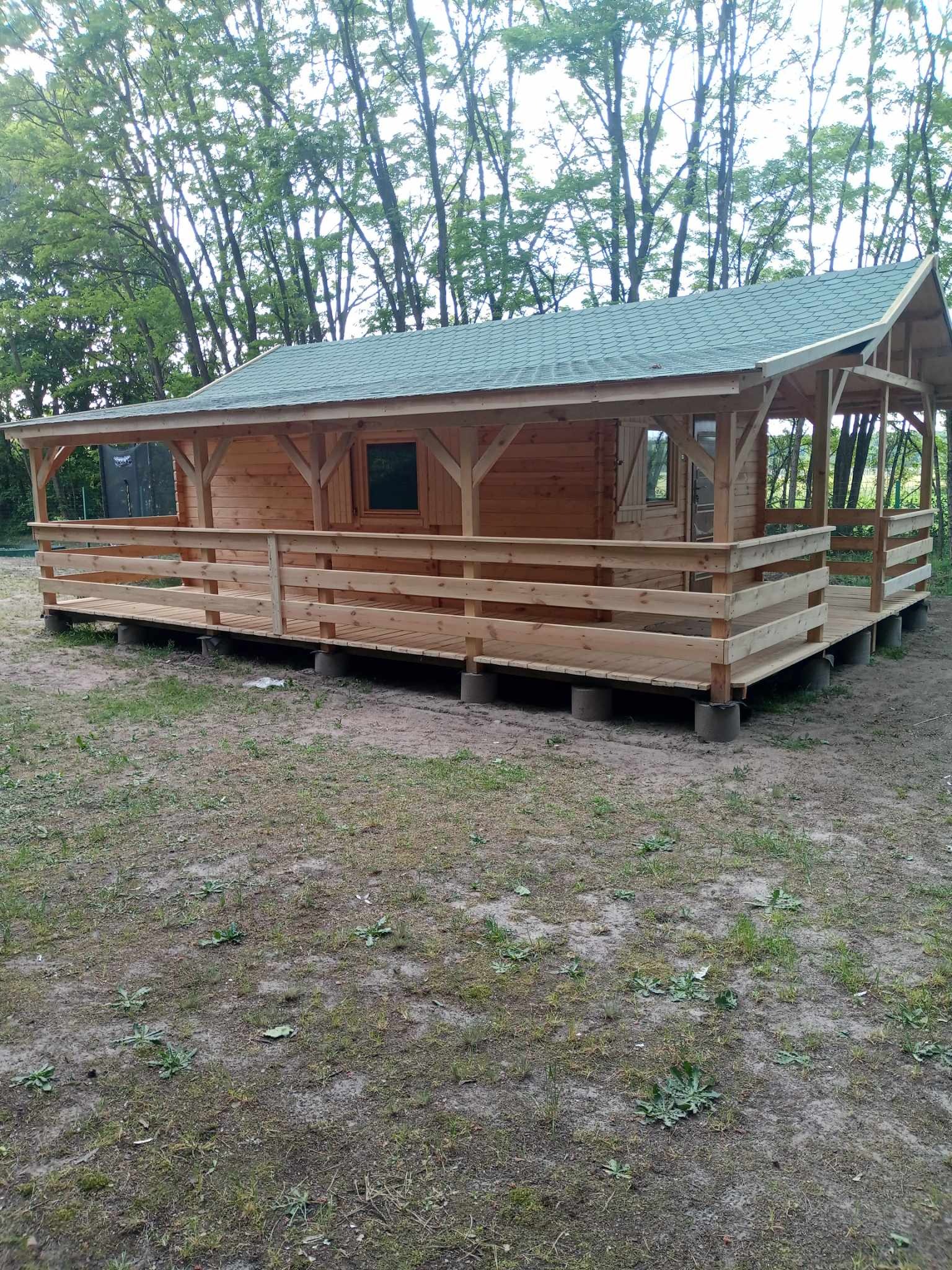Niewielki drewniany domek z zadaszonym tarasem, posadowiony na betonowych bloczkach, z zielonym dachem, otoczony drzewami i trawiastym terenem.