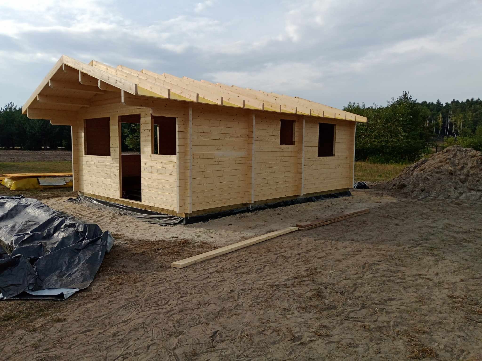 Wznoszona konstrukcja domu z drewna, widoczne ściany z bali i elementy więźby dachowej, teren budowy z piaskiem i folią ochronną.