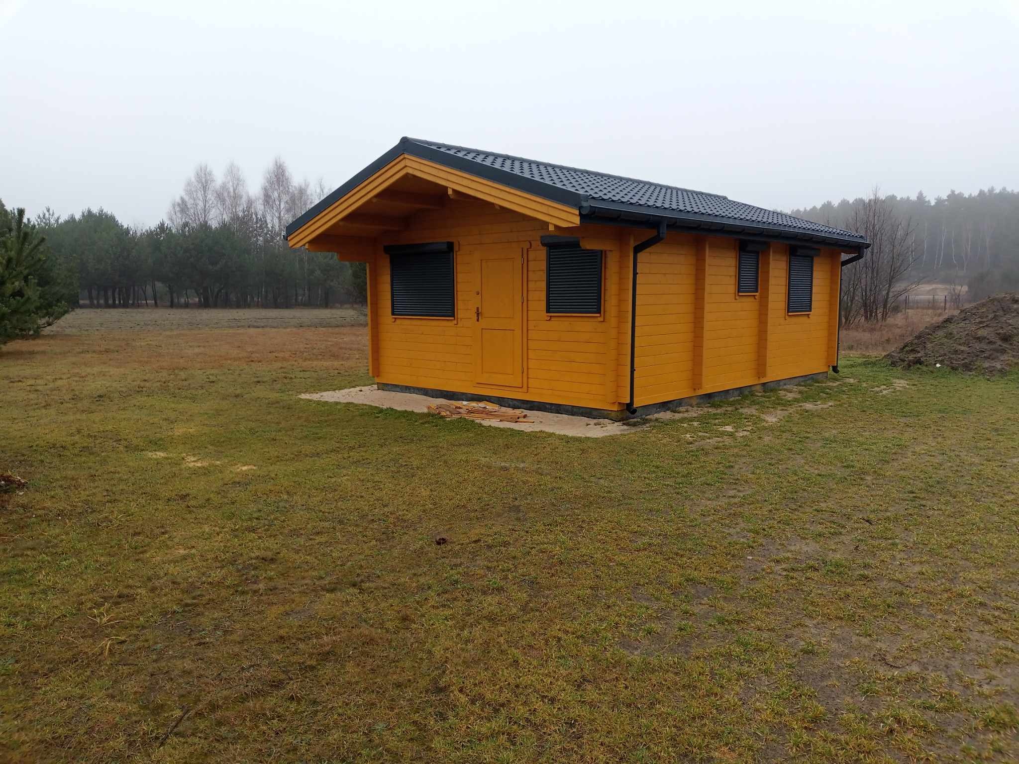 Żółty domek z ciemnym dachem i roletami zewnętrznymi, ustawiony na trawniku w pochmurny dzień, z lasem w tle.