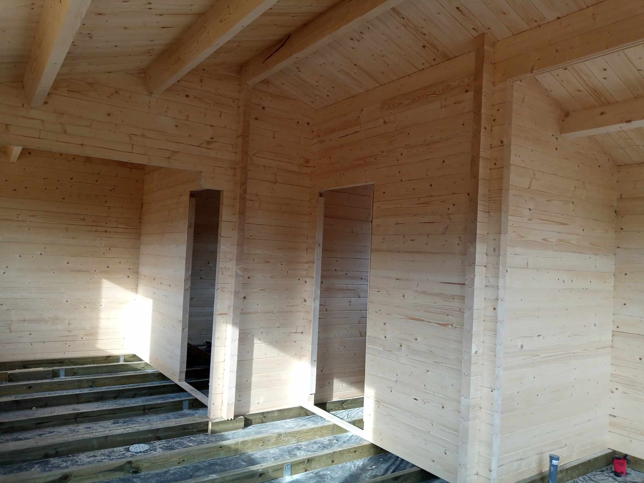 Wnętrze surowego drewnianego domu w trakcie budowy, widoczne ściany z bali, drewniany strop i przygotowane podłoże pod podłogę.