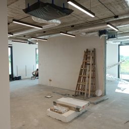 InstElectPro Andrii Dovhan - Wnętrze w stanie deweloperskim z widocznymi instalacjami elektrycznymi pod sufitem, liniowymi lampami LED, rozłożonymi przewodami w podłodze, drabina oparta o ścianę, oraz pracownik wykonujący...