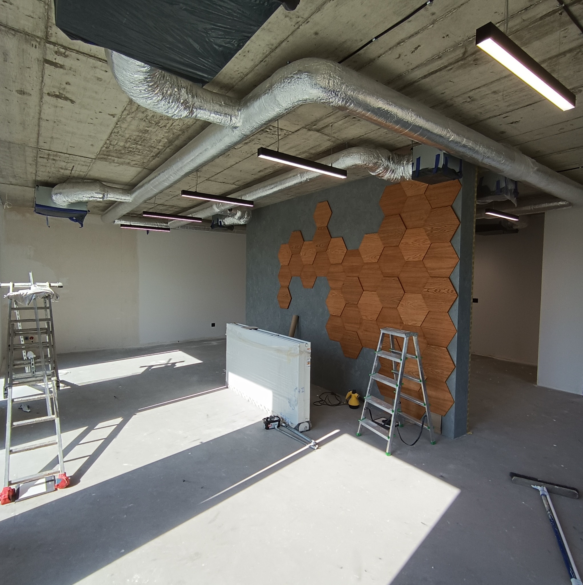 Wnętrze w trakcie wykańczania: widoczne rury wentylacyjne pod sufitem, oświetlenie liniowe, ściana z drewnianymi heksagonami, drabiny i narzędzia na podłodze.