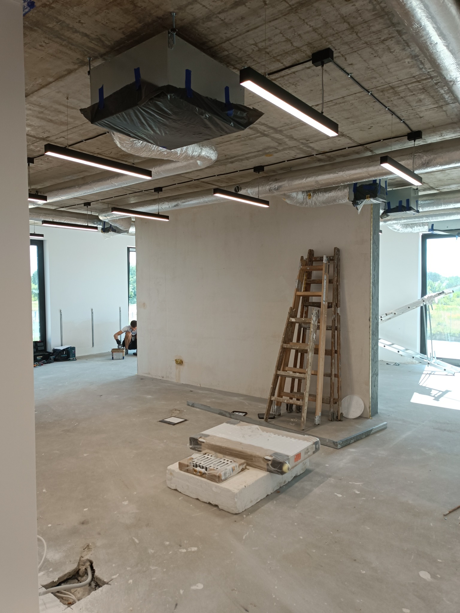 Wnętrze w stanie deweloperskim z widocznymi instalacjami elektrycznymi pod sufitem, liniowymi lampami LED, rozłożonymi przewodami w podłodze, drabina oparta o ścianę, oraz pracownik wykonujący...