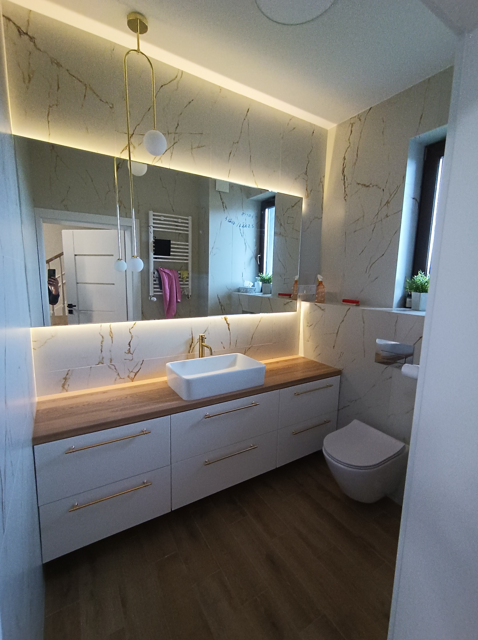 Nowoczesna łazienka z marmurowymi płytkami, podświetlanym lustrem nad drewnianym blatem z białą umywalką, złotą armaturą i wiszącą lampą z białymi kulami