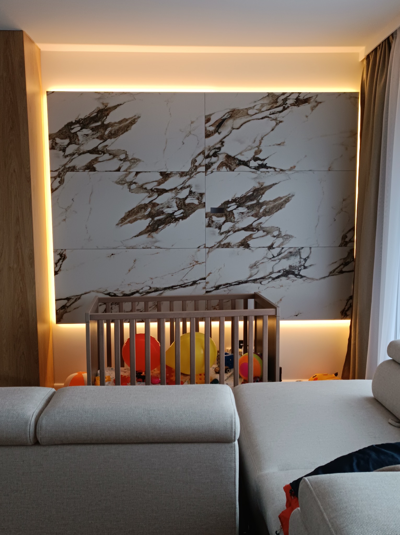 Podświetlana ściana z marmurowym wzorem za dziecięcym łóżeczkiem, widok zza sofy w salonie.