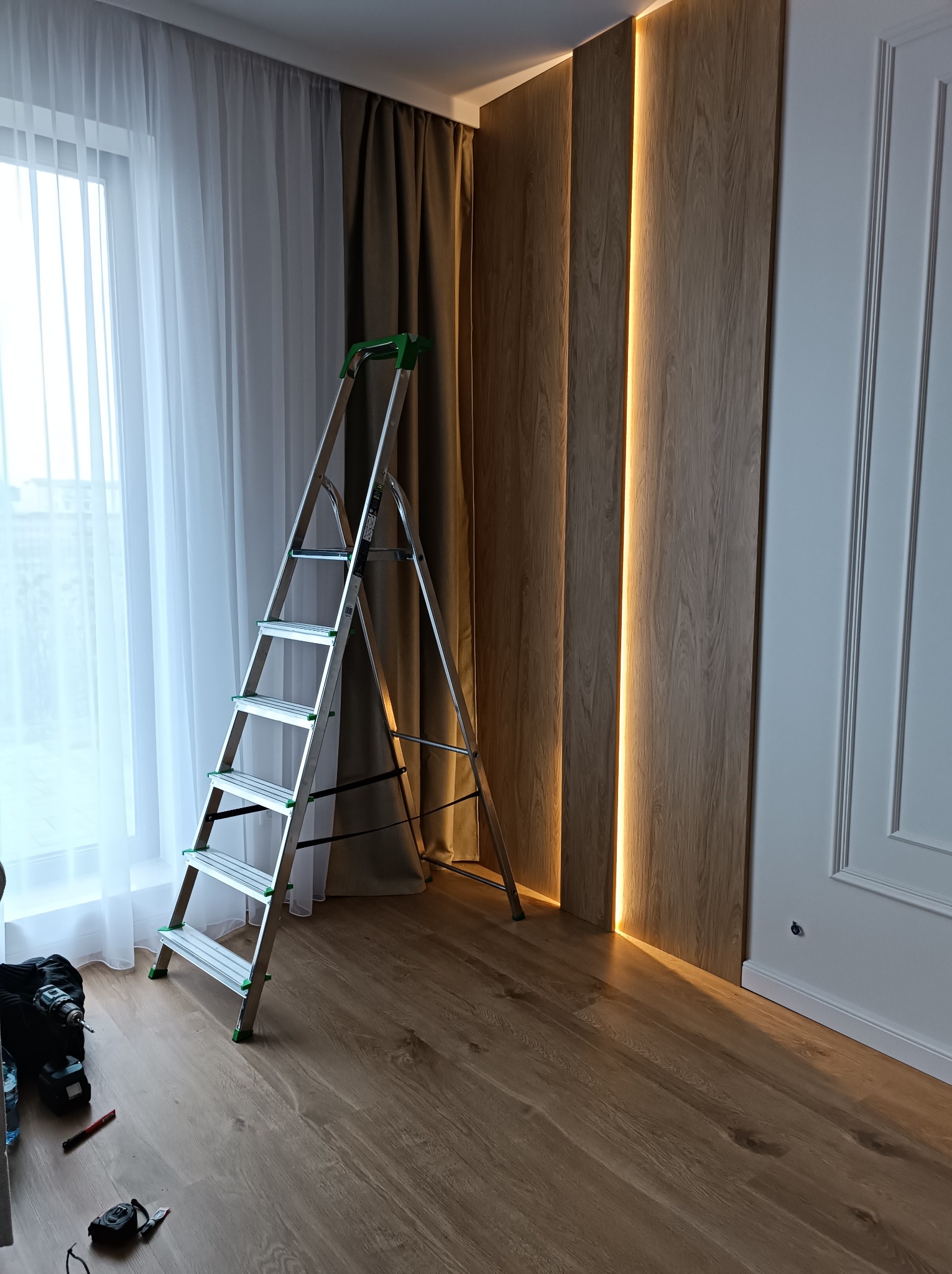 Wnętrze pokoju z drewnianą ścianą podświetloną paskami LED, drabina aluminiowa z zielonymi elementami oparta o ścianę, narzędzia i wkrętarka na podłodze.