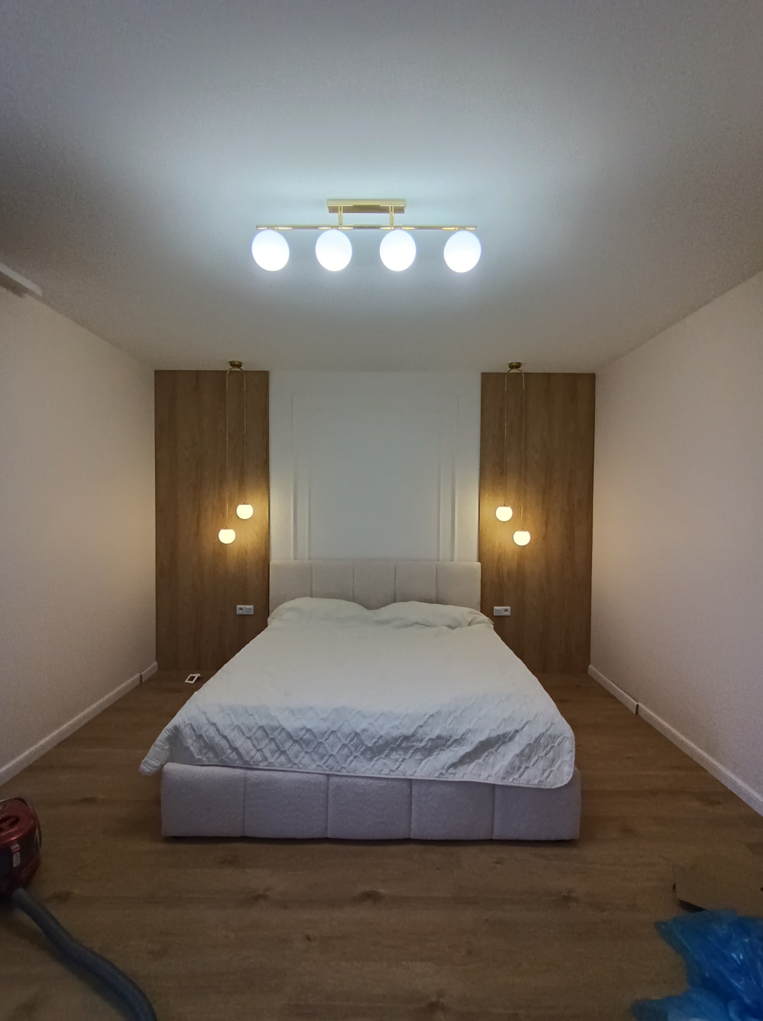 Nowoczesna sypialnia z podwójnym łóżkiem, drewnianymi panelami ściennymi i trzema punktami świetlnymi: centralnym plafonem i dwoma wiszącymi lampami po bokach łóżka.