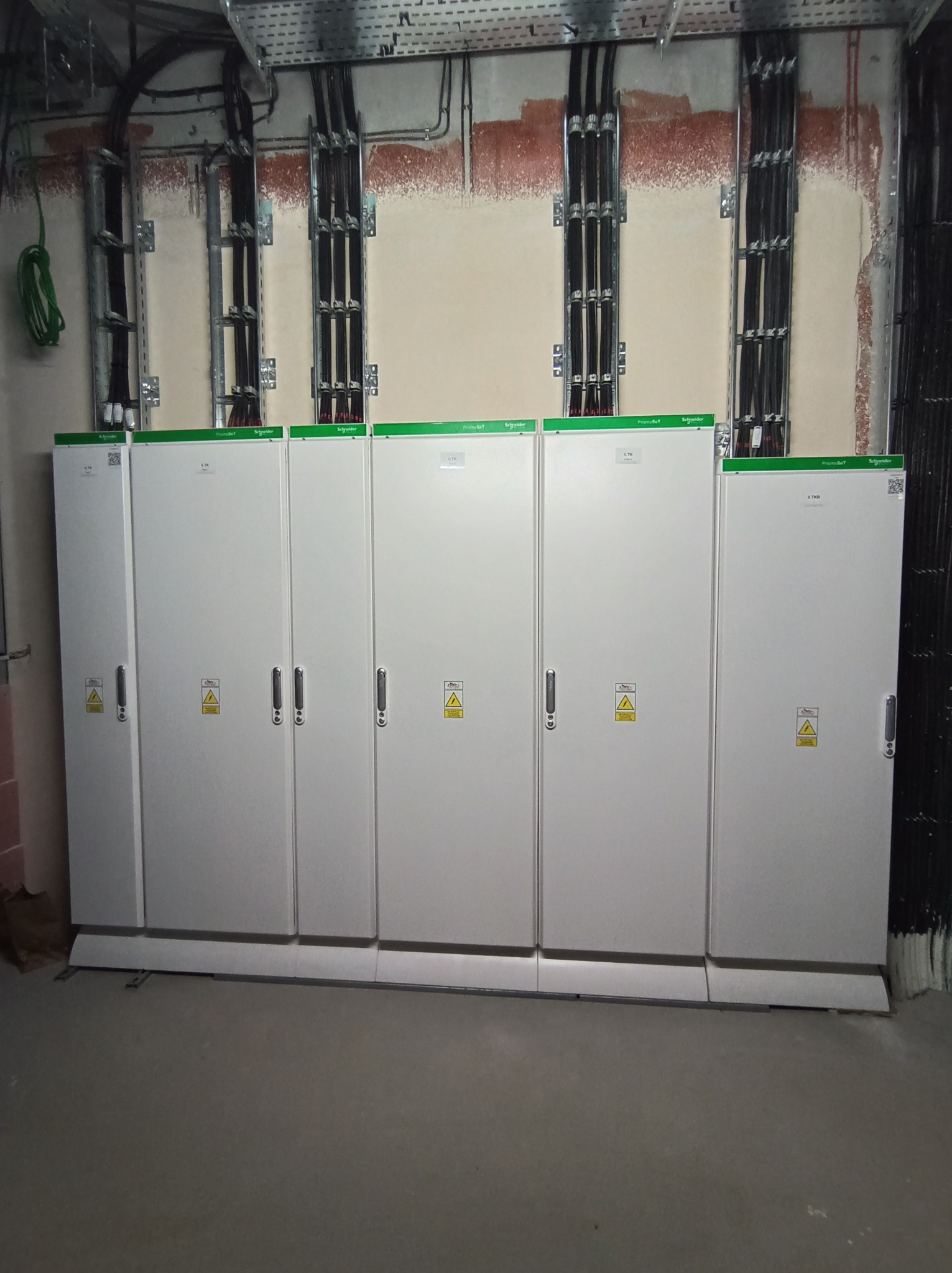 Rząd białych szaf elektrycznych Schneider PrismaSeT z żółtymi ostrzeżeniami, umieszczonych wzdłuż ściany z widocznymi kablami i metalowymi korytkami kablowymi pod sufitem.