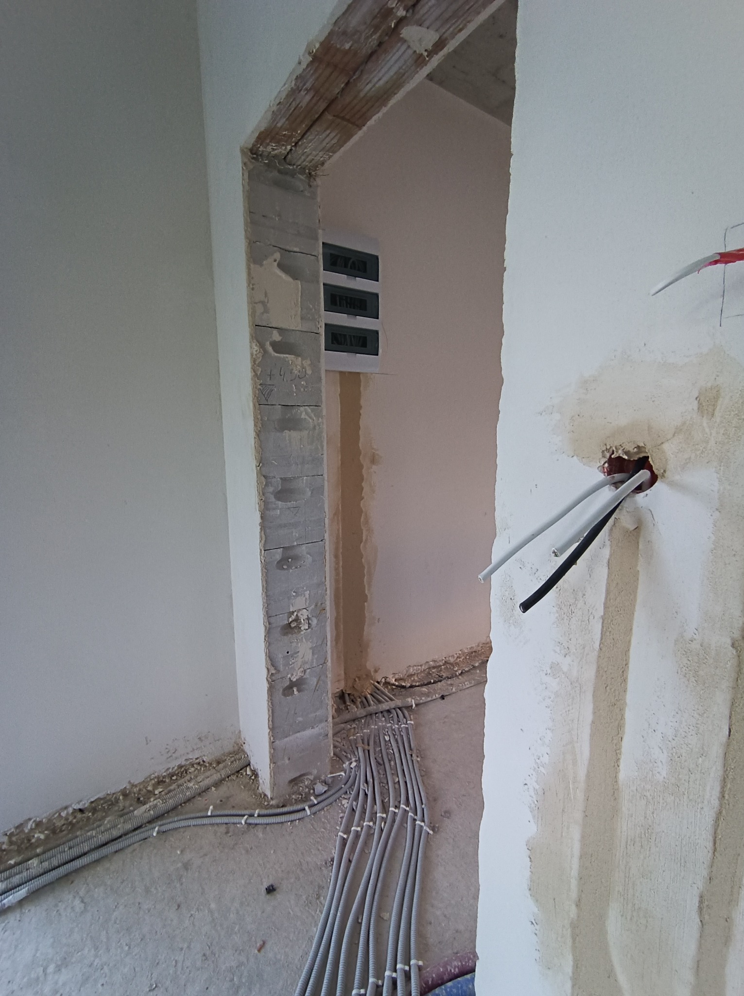 Wnętrze w trakcie remontu z widoczną rozdzielnicą elektryczną, kable w peszlach ułożone wzdłuż ściany oraz wystające przewody ze ściany w przygotowanym otworze.