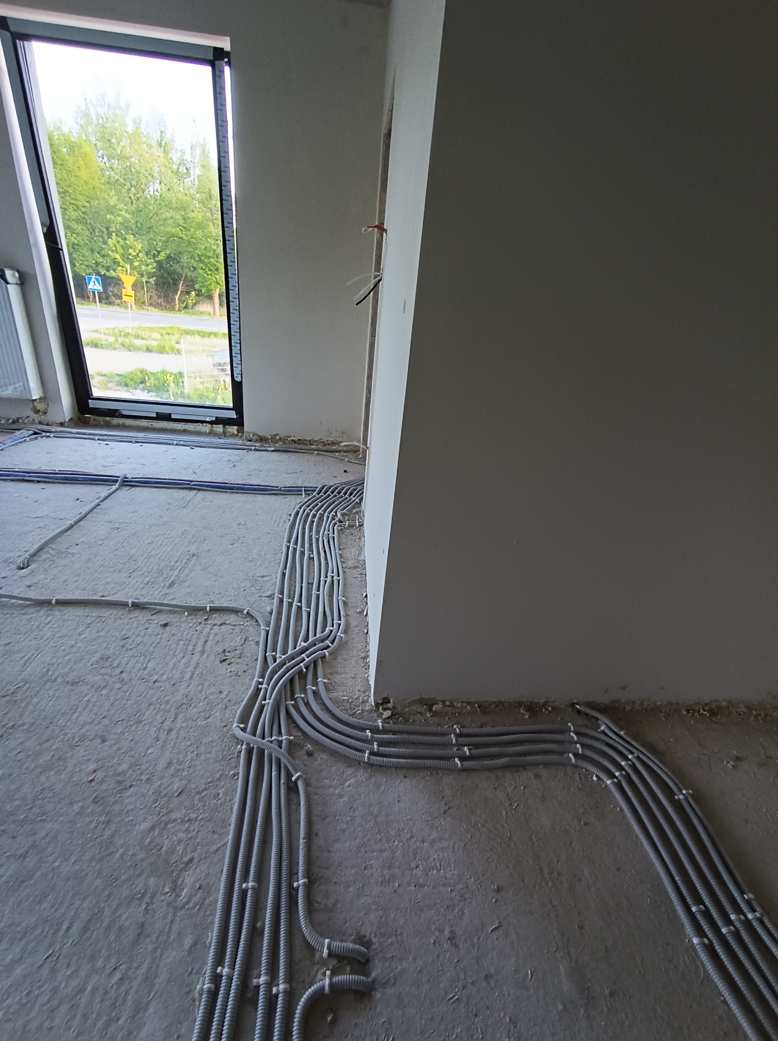 Szare, karbowane rury ochronne na przewody elektryczne ułożone równolegle na surowej posadzce w nowym budynku, widok z boku na okno.