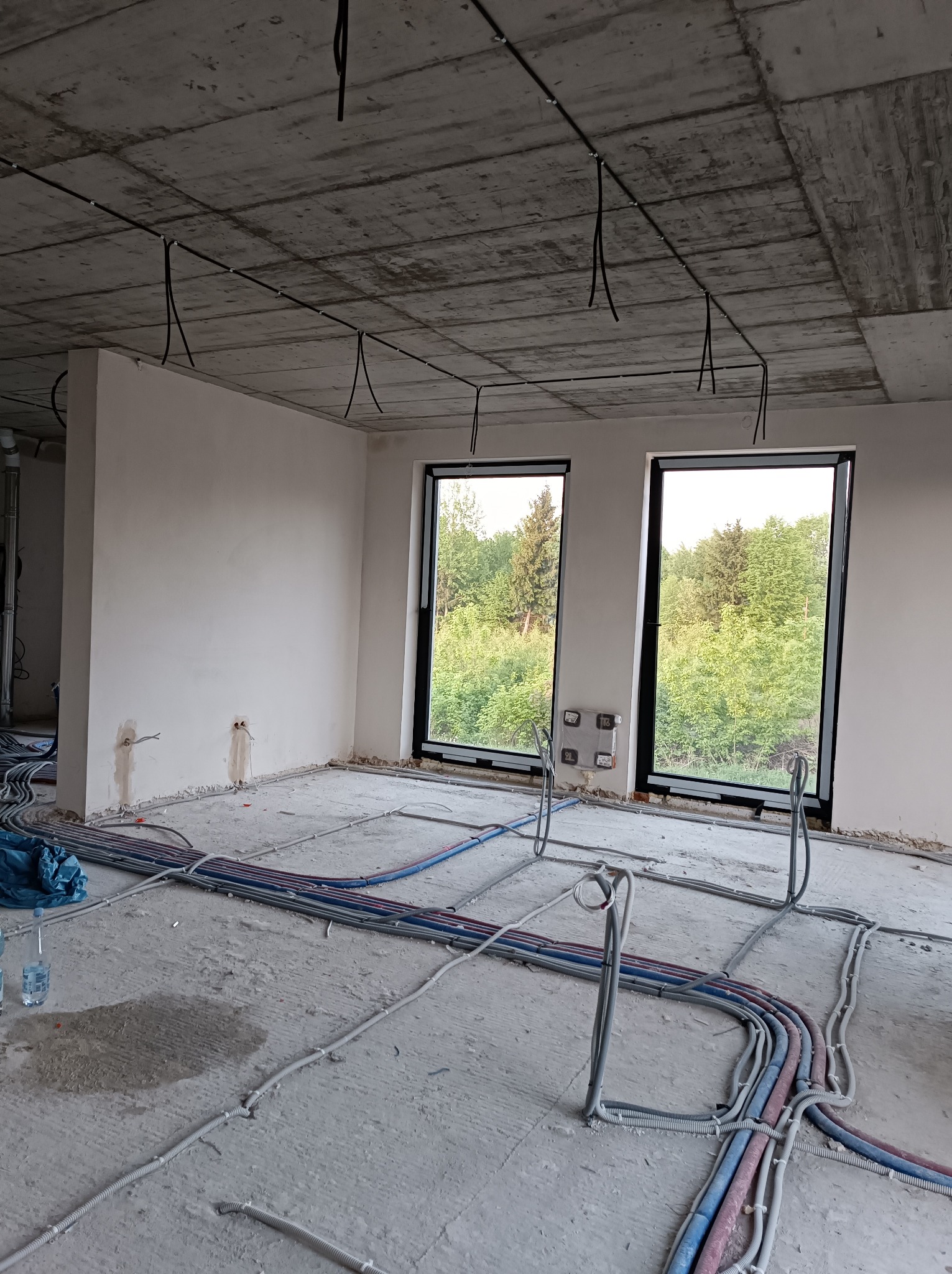 Wnętrze w stanie surowym z widocznymi kablami elektrycznymi prowadzonymi po podłodze i pod sufitem, dwa okna z widokiem na zieleń na zewnątrz.